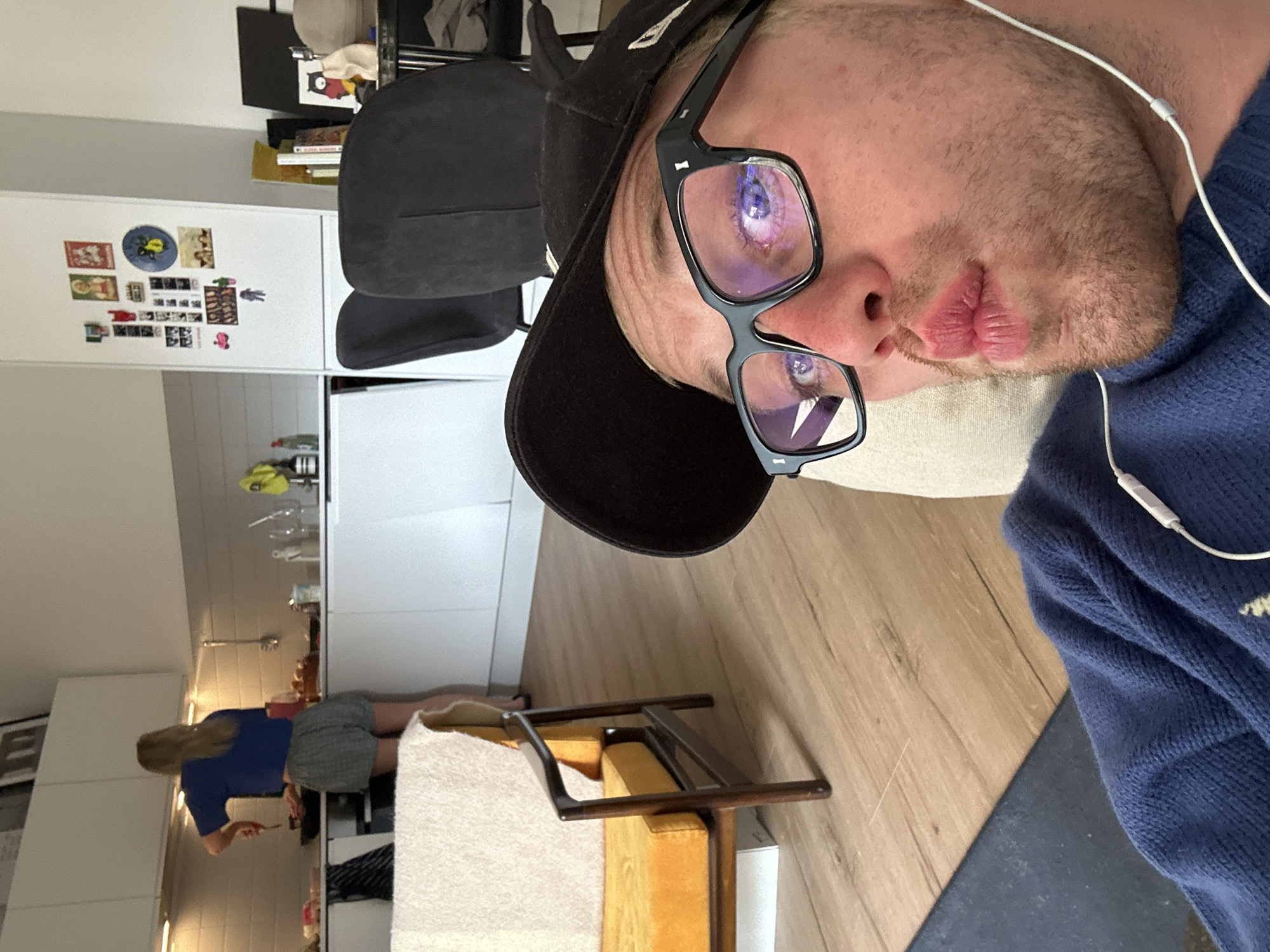 A selfie photograph depicts a man wearing a black baseball cap and glasses, with earbuds in his ears. He is looking at the camera with pursed lips. The background shows a domestic interior. In the distance, a woman with blonde hair is standing at a kitchen counter. The setting appears to be a modern apartment with white cabinets and appliances. There is no information within the image to place it in London, United Kingdom.