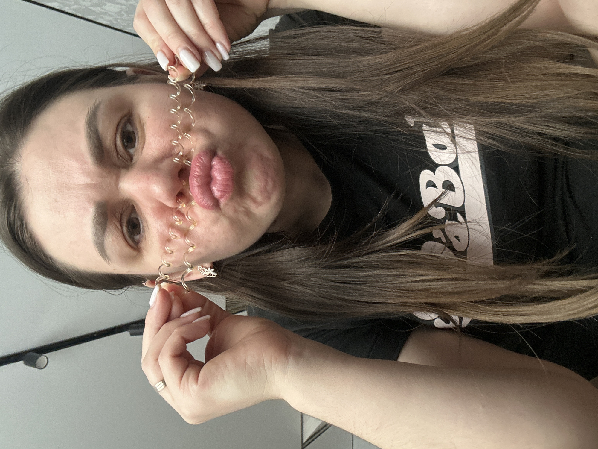 A woman with long, brown hair is holding up a gold-colored piece of jewelry with small, circular elements in front of her face. She has light skin, brown eyes, and is wearing a black t-shirt with a white design. Her lips are pursed. She has white nail polish on her nails and is wearing a ring on one finger. The background appears to be an interior space with a white wall and a black light fixture. The exact location within the city of Chaikovsky, Russia, cannot be determined from the image.
