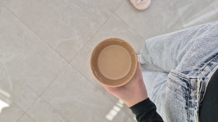 The image displays a downward-angled perspective of a person seated indoors in Batumi, Georgia. A pale-skinned left hand, extending from a dark long-sleeved garment, holds a light brown disposable paper cup. The cup contains an opaque, light brown liquid. The person's lap and upper thighs are visible, clad in light blue denim jeans with visible stitching and a metal rivet detail. Below the jeans, striped socks are faintly discernible on the left foot. A partial view of a light pink shoe or slipper is present in the lower right corner of the frame. The floor consists of light gray tiles with a subtle texture and visible diagonal grout lines. Strong, directional lighting from an unseen source casts distinct rectangular shadow patterns onto the tiled floor in the upper left quadrant.
