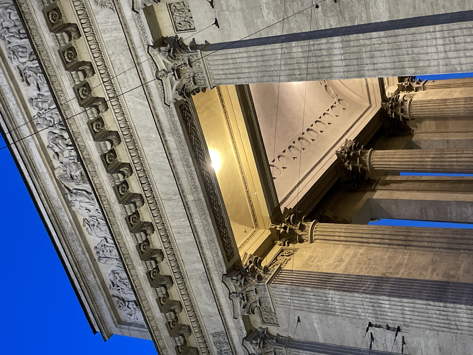 The image presents an upward, tilted view of the richly decorated facade of a neoclassical building, captured during dusk or night in Saint Petersburg, Russia. A striking deep blue sky forms the backdrop to the illuminated architectural elements.
Prominently featured are several monumental, fluted columns, topped with elaborate Corinthian capitals. Above these columns, an intricately carved entablature is visible. The frieze section boasts detailed bas-relief sculptures depicting numerous figures in dynamic poses, suggesting historical or mythological narratives. Further decorative elements, including a series of square corbels adorned with rosette motifs, are visible along the underside of the cornice.
Artificial lighting, likely from recessed fixtures or the building's interior, casts a warm, golden glow on the ceiling or soffit, revealing geometric patterns and highlighting the textures of the stonework. This illumination creates strong contrasts and shadows that emphasize the depth and complexity of the architectural details. Thin wires or cables are present, stretching across the frame, which could be part of the building's lighting system or urban infrastructure. The overall impression conveyed is one of immense grandeur, historical artistry, and architectural sophistication, characteristic of the city's renowned structures. No discernible text is visible within the frame.