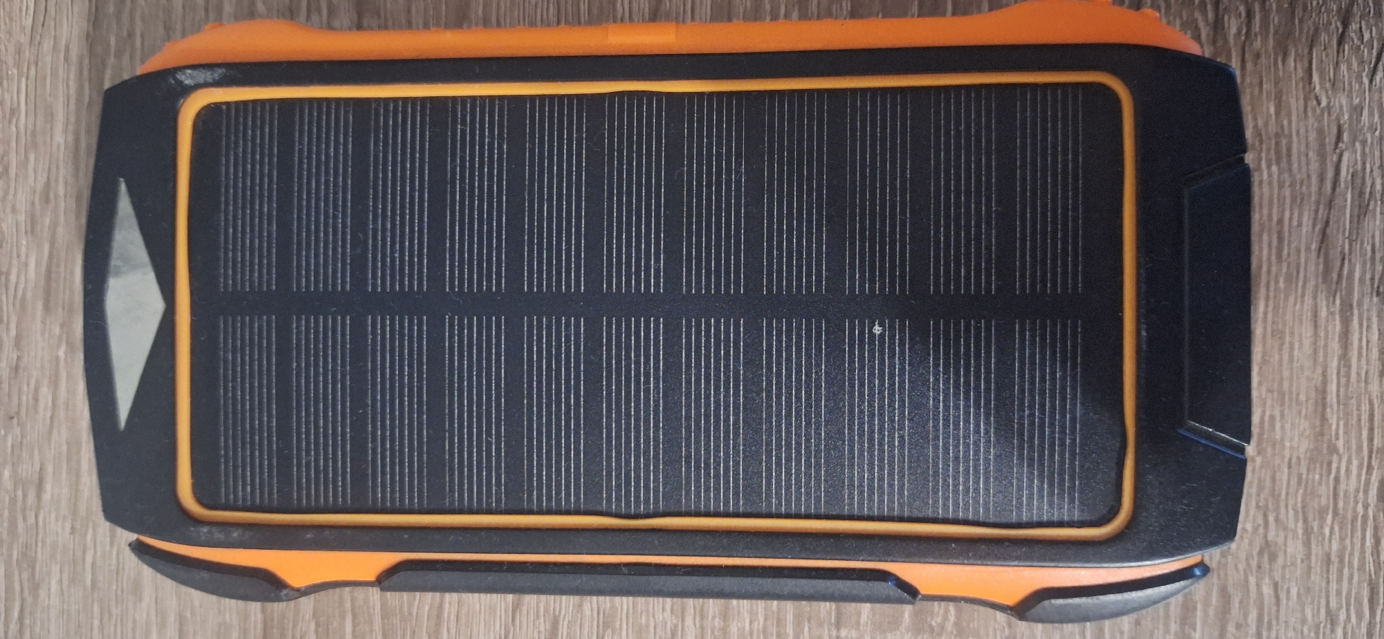 This is a top-down, medium shot of a solar power bank resting on a wooden surface. The power bank is predominantly black with orange accents around its edges and a textured orange material on its top and bottom. The central feature of the device is a rectangular solar panel, composed of numerous vertical black photovoltaic cells with thin silver lines running between them. The solar panel is bordered by a thin, bright orange trim. On the left side of the power bank, there is a triangular, light gray marking.
The wooden surface appears to be a table or countertop, with visible grain patterns and a light brown color. The lighting suggests an indoor environment, possibly with ambient light from a window or overhead source, creating subtle shadows around the power bank. No people are visible in the image. The overall impression is of a functional electronic device, likely intended for outdoor or emergency use, placed on a simple, neutral background. No text is discernible on the power bank or the surrounding surface. The image is static and focuses solely on the object itself.
