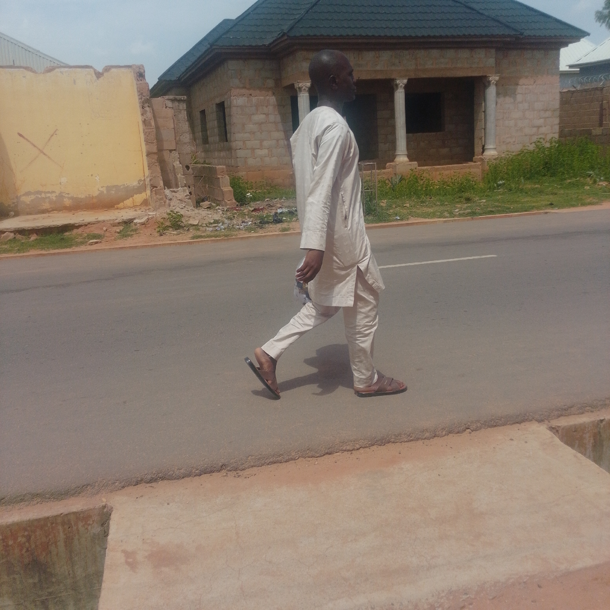 The image depicts a man walking on a paved road in Gombe, Nigeria. The man is wearing a light-colored traditional outfit and sandals. He is walking from left to right, facing away from the camera. In the background, there is a building under construction, with exposed bricks and pillars. To the left of the building, a partially demolished yellow wall is visible. The sky is overcast. The road appears to be in a somewhat neglected state, with some debris on the side. The environment suggests an urban or semi-urban setting within Gombe.