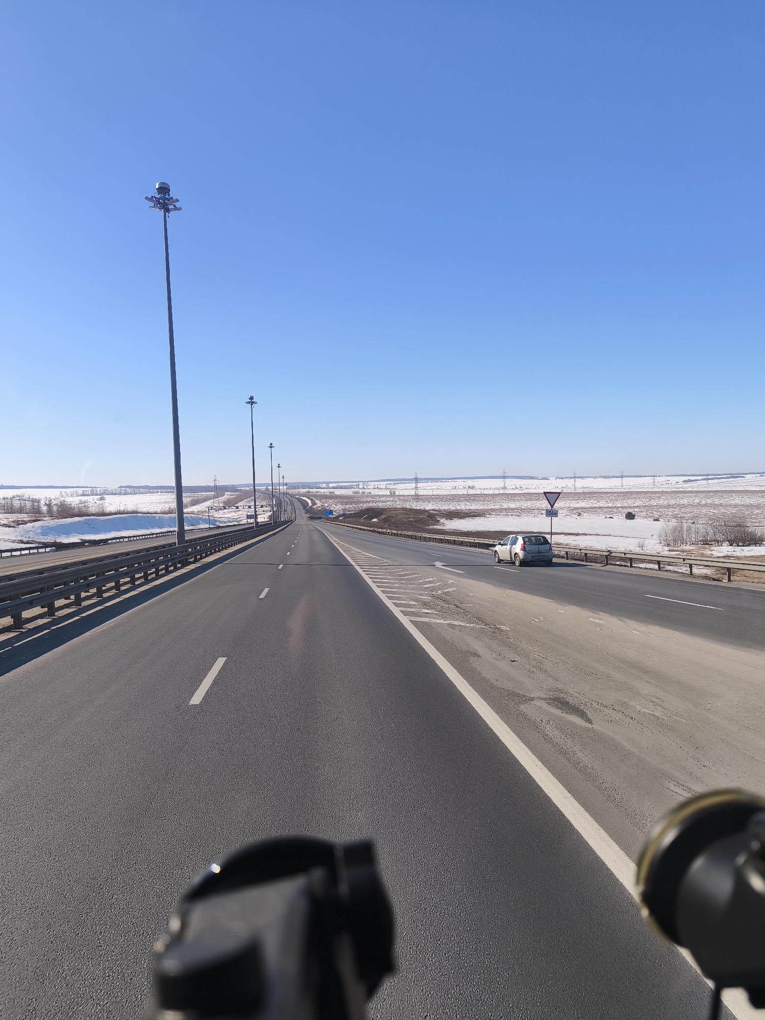The image depicts a multi-lane highway bridge under a clear blue sky, likely during daytime. The landscape on either side of the bridge is flat, predominantly snow-covered, and contains power lines and sparse vegetation. The road surface is asphalt. Roadside safety barriers are visible along the edges of the bridge. A light-colored car is present on the right side of the road, partially obscured by a road sign indicating "Yield." The location is indicated as Shali, Russia. The bottom portion of the image contains the vague shape of a camera. There are no people present.