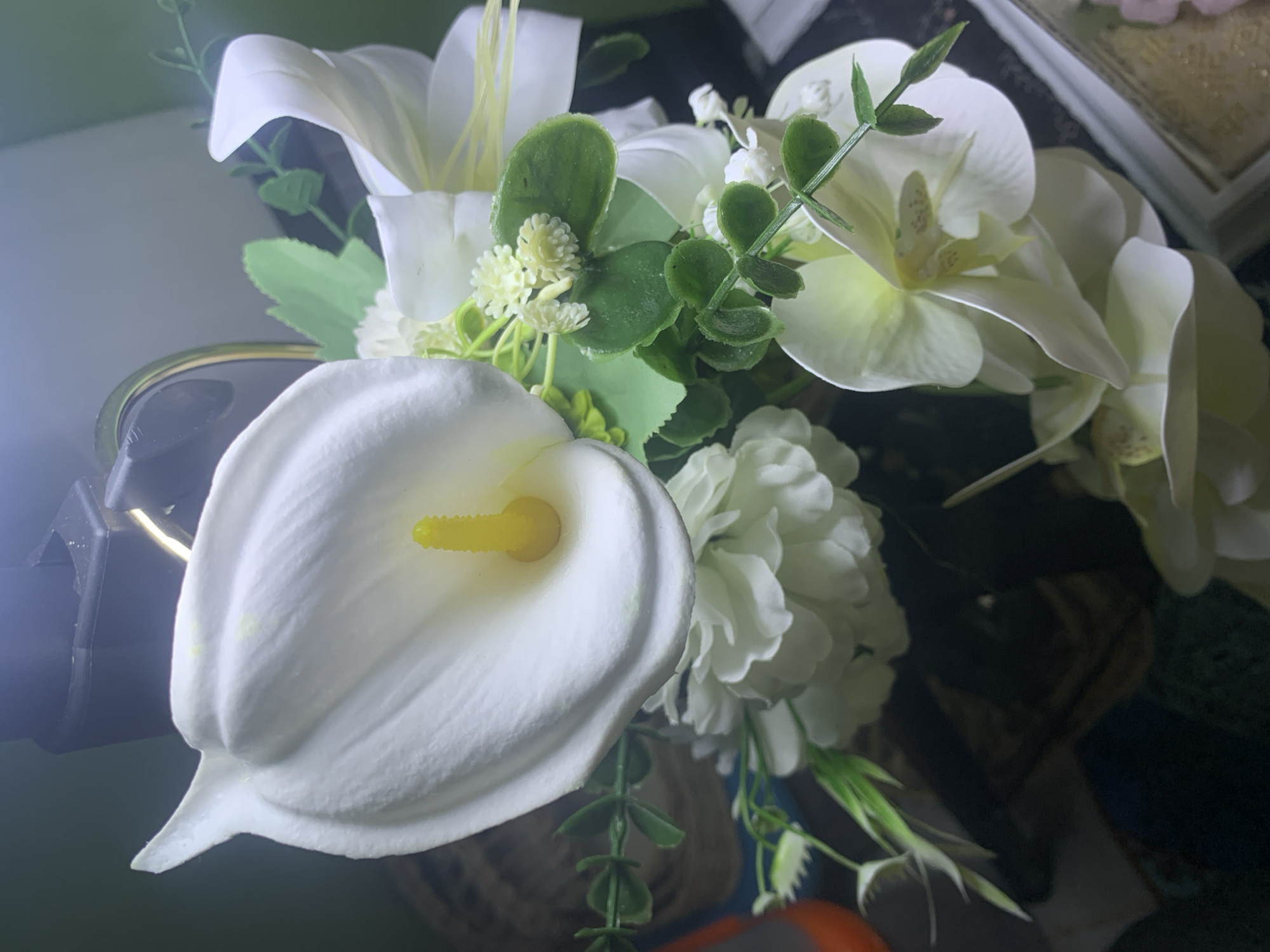 A close-up shot showcases a vibrant bouquet of artificial white flowers, including calla lilies, orchids, and other delicate blooms, nestled amongst lush green foliage. The arrangement is artfully composed, with the prominent calla lily, its creamy white spathe unfurling around a bright yellow spadix, taking center stage. Smaller, intricate white flowers and clusters of round green leaves add depth and texture to the bouquet. The backdrop is a mixture of dark, out-of-focus elements and lighter, softer tones, suggesting an indoor setting, possibly a home or event space. The lighting appears to be artificial, casting a slight glow on the petals and highlighting the details of the floral arrangement. There is no discernible activity or event taking place, and no visible text. The overall impression is one of artificial elegance and delicate beauty. The location context of Kuala Pembuang, Indonesia, does not provide any specific visual cues within the image itself.