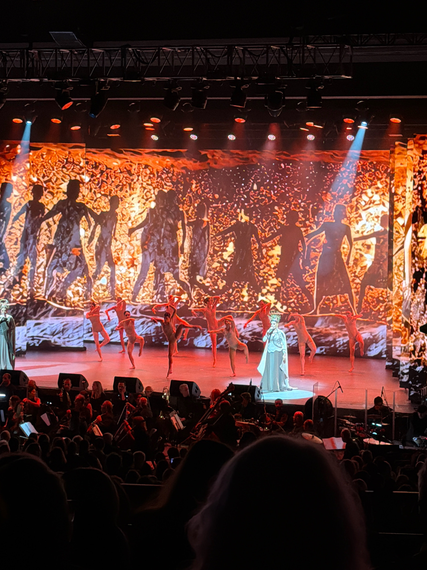 The image depicts a live performance taking place on a stage in Los Angeles, California, United States of America. The stage is illuminated with dramatic lighting, featuring a large projection screen displaying silhouetted figures against a fiery, abstract background. In the foreground, a group of dancers in light-colored costumes are performing various poses. Center stage, a performer in an elaborate white gown and crown stands, appearing to sing into a microphone. Behind the dancers and main performers, an orchestra is visible, with musicians playing instruments. The audience is seated in a dark, tiered theater, with the backs of their heads and shoulders visible in the foreground. The overall scene suggests a theatrical or musical production, possibly an opera or a ballet, given the costumes and staging.
