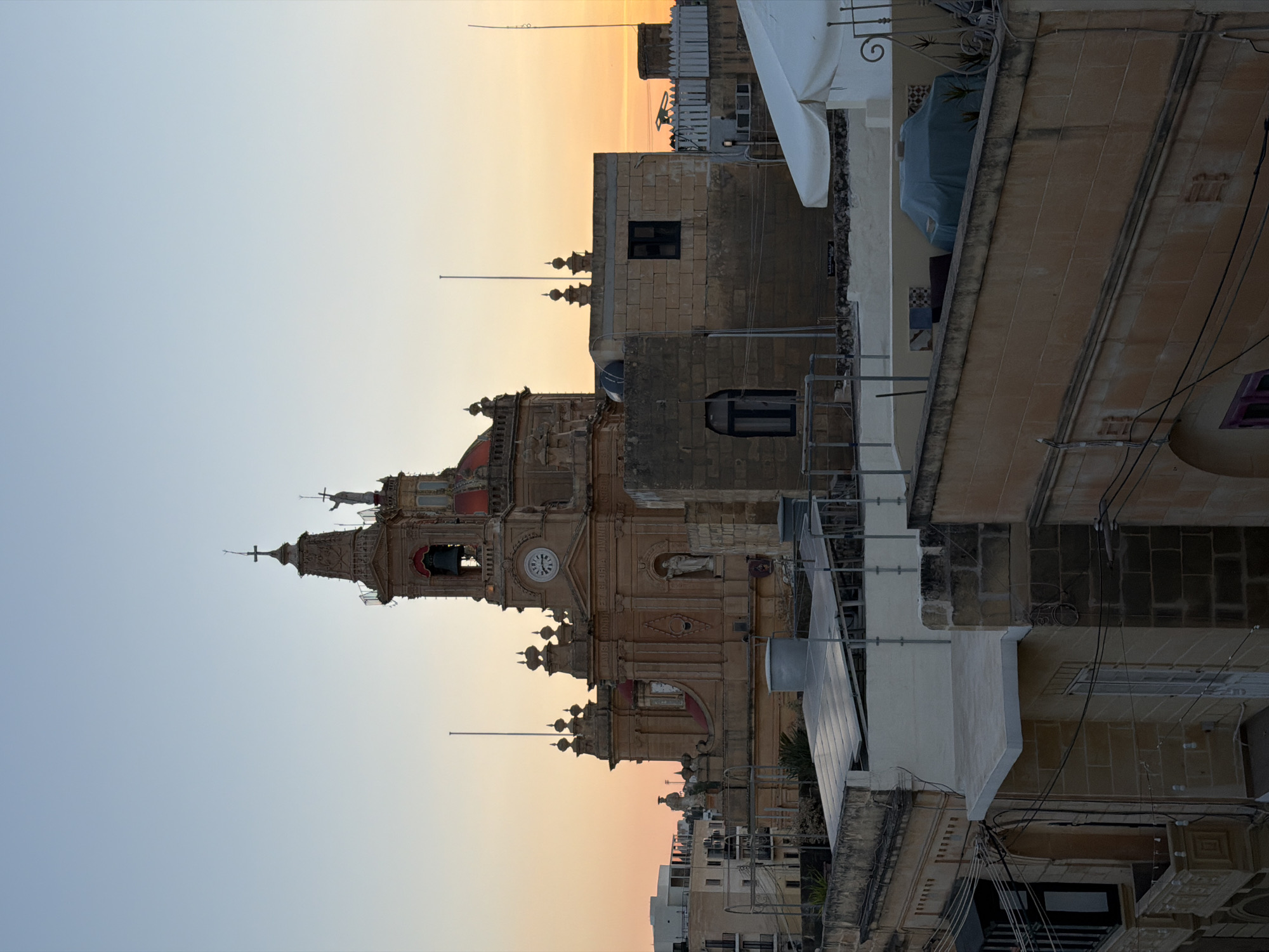 In Fontana, Malta, at sunset, a multi-story church with ornate architecture and a clock tower dominates the skyline. Several buildings with flat roofs, some featuring solar panels, are visible in the foreground, interspersed with residential structures and various rooftop elements. The sky transitions from a clear blue to a warm orange hue, casting a soft light over the scene.