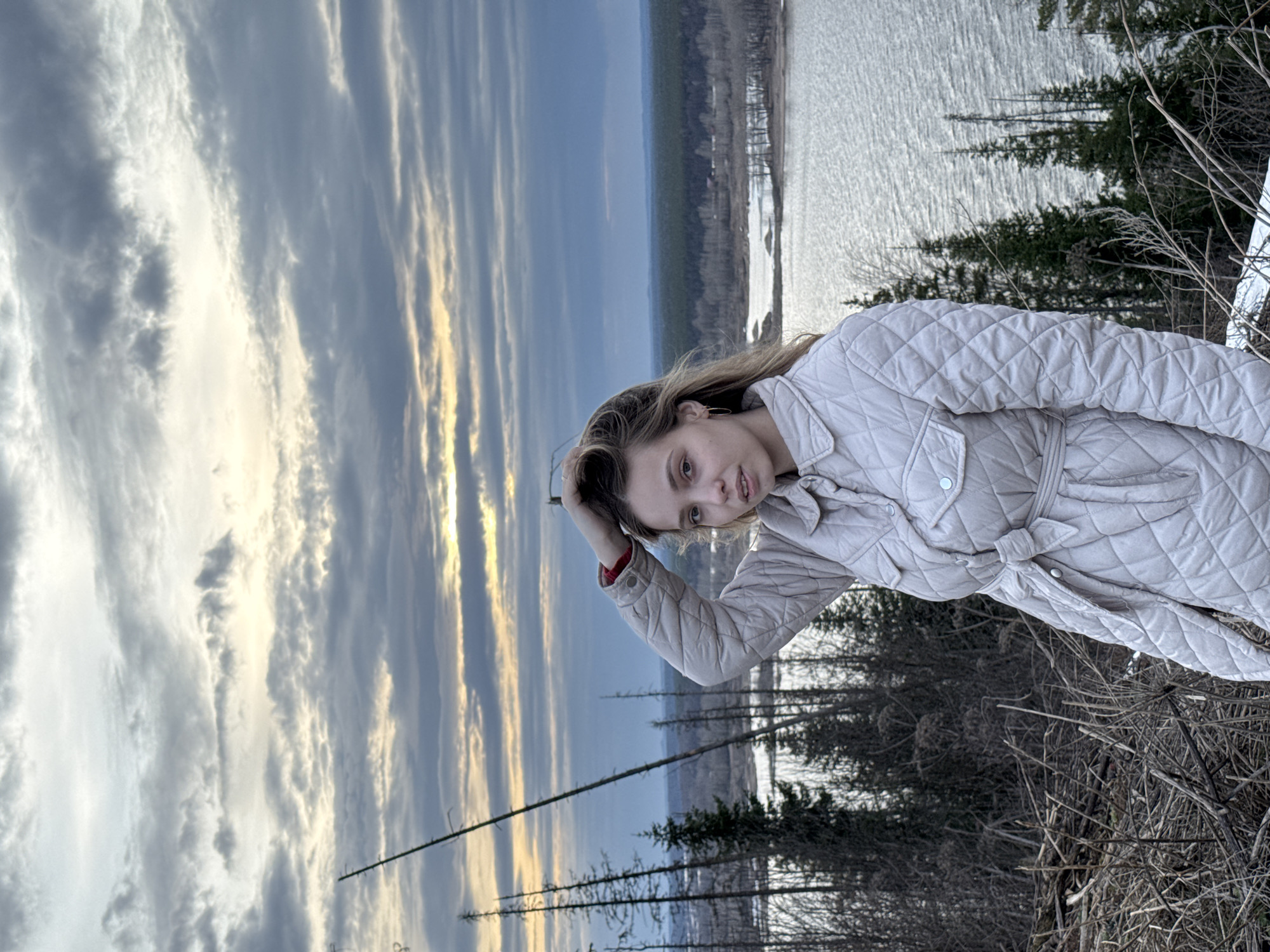 The image shows a woman standing outdoors, likely in a natural setting near a body of water. The location appears to be in or near Chaykovsky, Russia, based on the environment. The woman is wearing a light-colored, quilted jacket with a belt. The background includes a landscape with a body of water, trees, and a cloudy sky with some patches of light, suggesting either sunrise or sunset. The foreground includes some dry grass and vegetation.