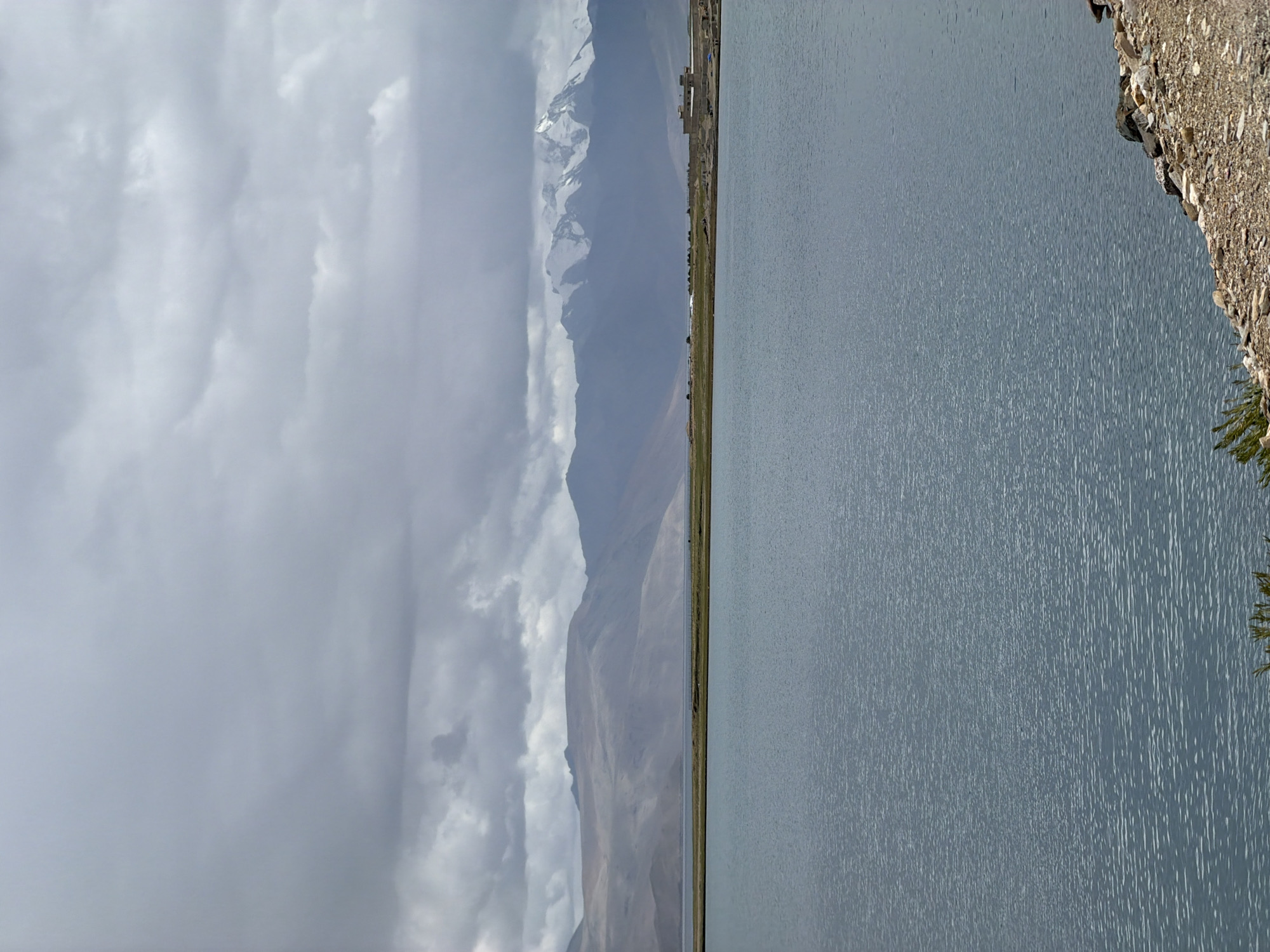 A wide shot captures a tranquil lake with a vast, overcast sky. The lake's surface is a muted blue, rippled by a gentle breeze, reflecting the cloudy heavens. In the background, a low, arid mountain range stretches across the horizon, its slopes a soft brown and beige. To the right, a section of the range is capped with snow-covered peaks, standing out starkly against the muted tones of the lower mountains.
Along the lake's far shore, a strip of green grass creates a distinct line between the water and the mountains. Further to the right, nestled against the foothills, is a cluster of buildings with light-colored walls, some appearing to be under construction or a small settlement. The immediate foreground shows a rocky, gravelly shoreline with sparse vegetation.
The scene is devoid of human presence, focusing entirely on the natural landscape. The location is identified as Karzok, India, a village situated on the banks of Tso Moriri lake. The image conveys a sense of vastness and quietude under a heavy, gray sky, with the snow-capped mountains hinting at the high-altitude, remote nature of the location.