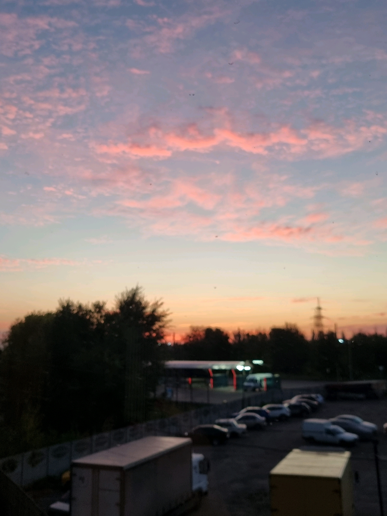 The image depicts a sky with pink and blue hues, indicative of a sunset. Below the sky, a line of trees is visible, silhouetted against the fading light. In the mid-ground, a parking lot is sparsely populated with several cars. Further in the background, a building with illuminated signs and a transmission tower are present. The overall scene appears to be an outdoor area within the city of Kryvyi Rih, Ukraine, possibly a commercial or industrial zone. There are no discernible individuals or specific actions taking place in the image.