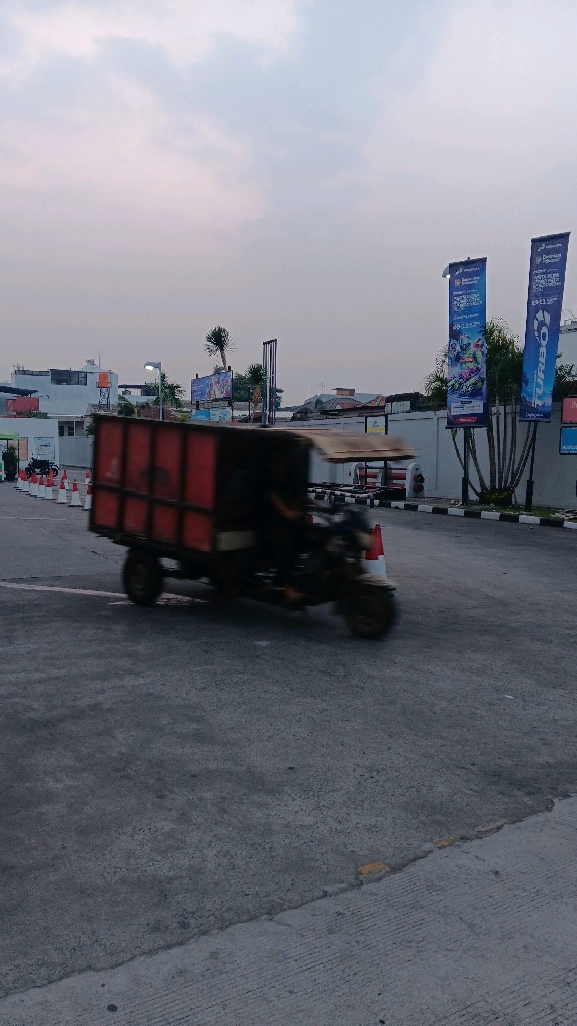 In South Jakarta, Indonesia, an outdoor scene captures a blurred motion of a three-wheeled truck with a red cargo box driving on a paved road. The truck is in the foreground, its movement creating a motion blur effect. Behind the truck, a row of orange traffic cones lines the edge of the road. In the background, there are buildings, palm trees, and several large banners. The sky is overcast with a soft gray hue. The overall impression is of a typical street scene in an urban environment.