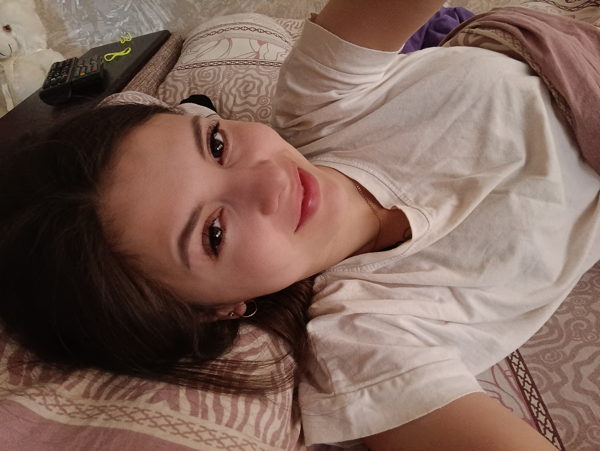 A young woman with long, dark hair is lying on a bed in what appears to be an indoor setting. She is wearing a white T-shirt. The bedding has a patterned design. A remote control and a set of yellow hair ties are visible on a surface adjacent to the bed. The presence of the remote control and the woman's relaxed posture suggest a domestic environment. The image does not contain any information regarding the city of Chaikovsky, Russia.