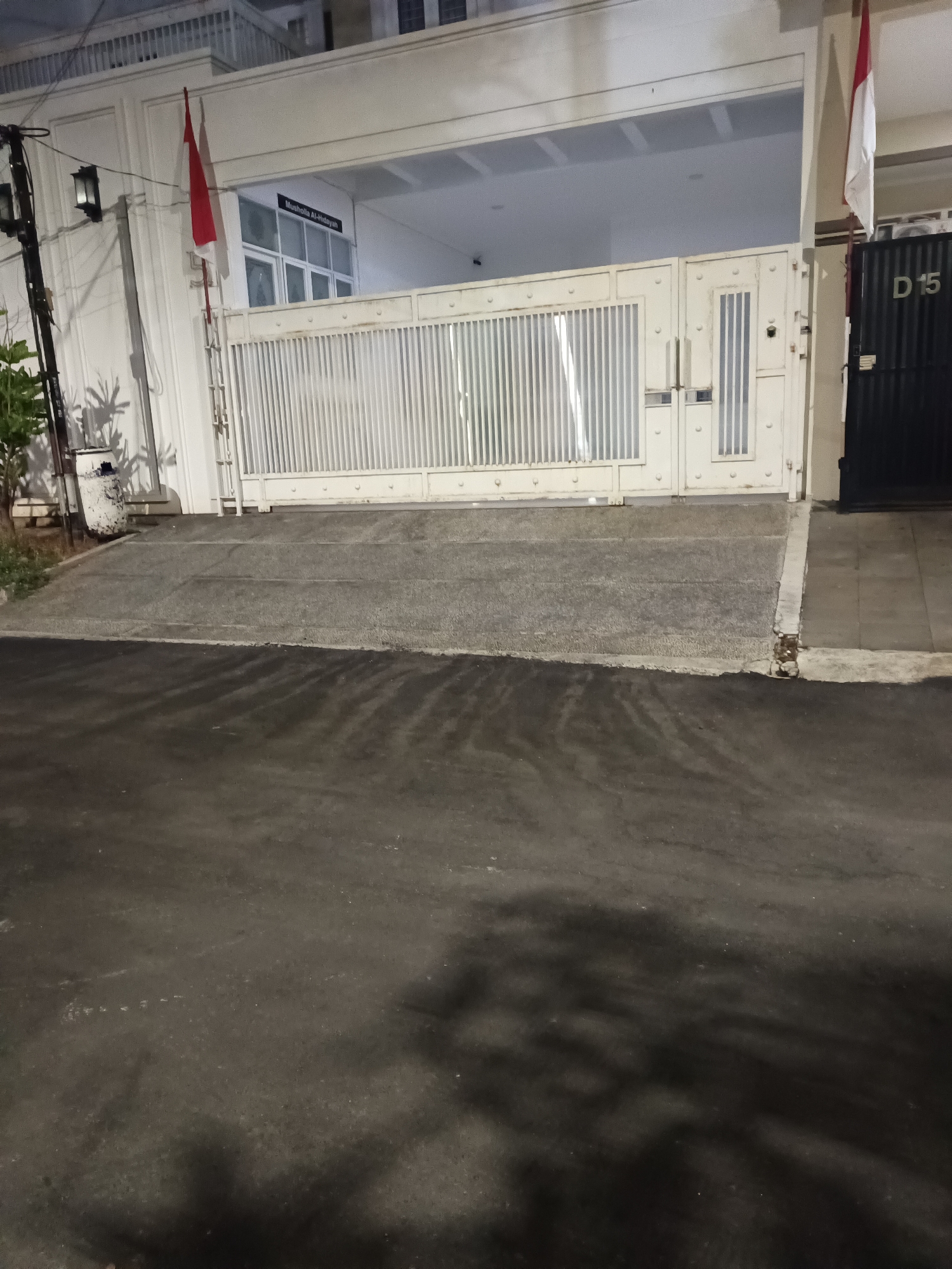 The image shows the entrance of a building with a white fence and a black gate on the right. The building appears to be a mosque or a prayer hall, as indicated by the sign that reads "Musholla Al-Hidayah". There are two Indonesian flags displayed on poles at the entrance. The street in front of the building is paved with asphalt and concrete. The scene is dimly lit, suggesting it is nighttime or early morning. There are no people visible in the image. The building is located in North Jakarta, Indonesia.