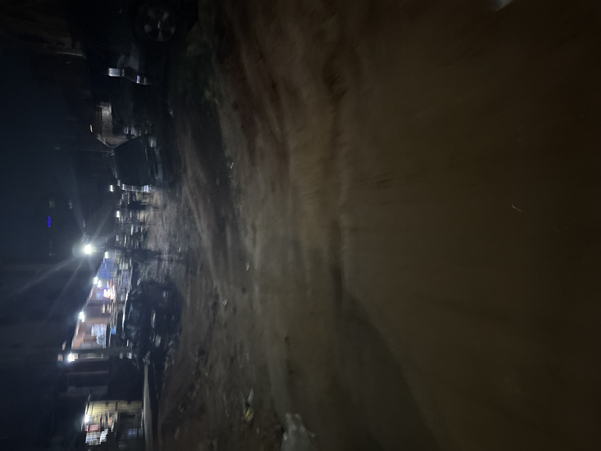 The image depicts a dimly lit, unpaved street in Onitsha, Nigeria, at night. The ground is uneven and appears to be made of dirt and gravel, with evidence of water accumulation or puddles. Several dark-colored vehicles are parked or driving on the street, some with their headlights illuminated. Buildings with lit windows are visible on the left side of the street, suggesting commercial or residential structures. In the background, figures of people are indistinct due to the low light and motion blur, indicating movement or activity. The overall atmosphere is gritty and urban, characteristic of many parts of Onitsha.