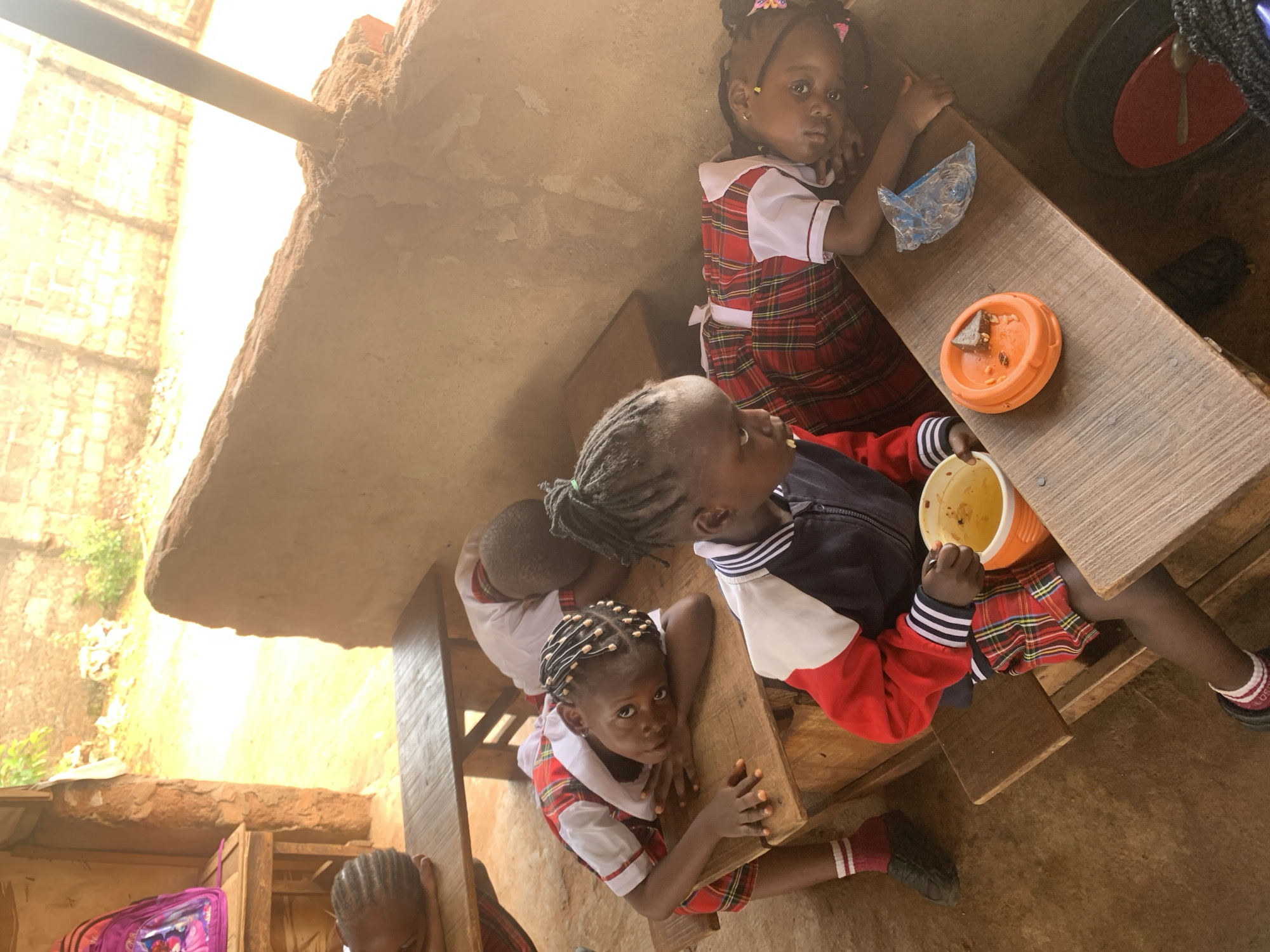 In Ukwu-Abwa, Nigeria, a group of young children, likely students, are seated at wooden tables eating. They are wearing plaid uniforms with white collars and sleeves and what appear to be red sweaters or jackets. One child holds a bright orange bowl. An orange plate with remnants of food sits on the table in front of another child. A blue surgical mask lies discarded on the table nearby. A pink backpack sits beneath a table. The setting appears to be an open-air classroom or eating area with rough-hewn walls.