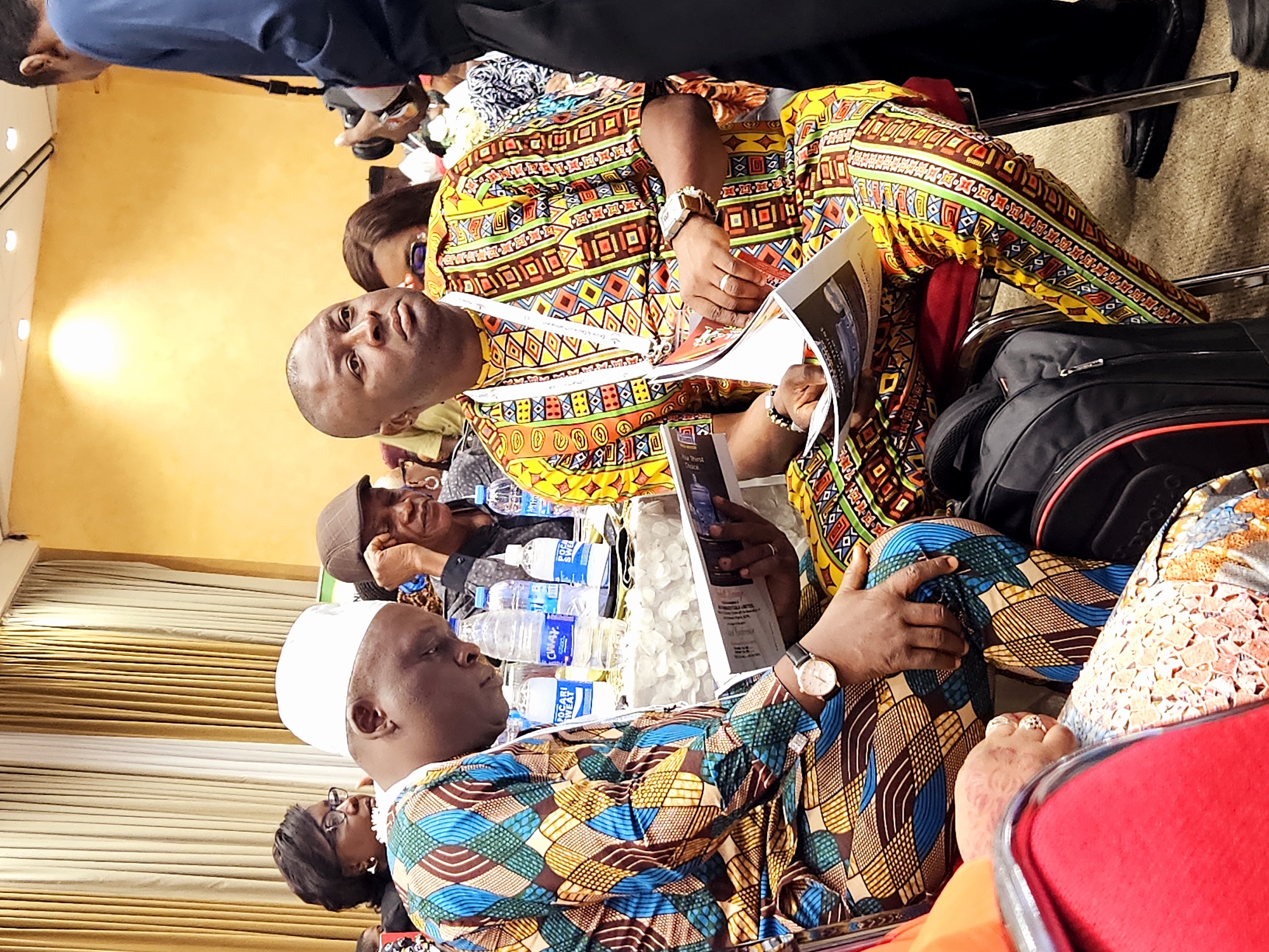 The image depicts an indoor event, likely a conference or meeting, taking place at the International Conference Centre in Abuja, Nigeria. Two men, dressed in vibrantly colored African attire, are seated in the foreground. The man on the left wears a white skullcap and a patterned dashiki with geometric designs in blue, brown, green, and yellow. He is looking down at a document he is holding in his hands. On the right, another man, also in a colorful dashiki featuring orange, yellow, red, green, and black patterns, is holding a magazine. He is looking forward, and a wristwatch is visible on his left wrist. A partially visible individual in a dark blue shirt is to the right of the frame, holding a camera. In the background, other attendees are seated, with a woman in glasses visible on the far left. The setting includes draped curtains in the background and tables with water bottles.