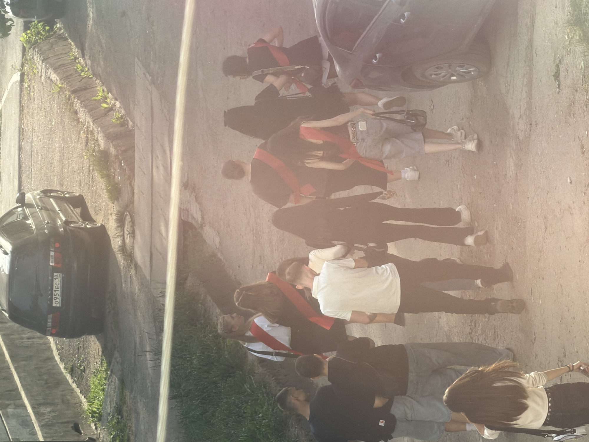 The image depicts a group of people gathered on a paved surface next to a road. Most of them are wearing graduation sashes. There are several parked cars nearby. One car’s license plate reads “O951EC 18”, suggesting the location is in the city of Votkinsk, Russia, part of the Udmurt Republic (region code 18). The environment appears to be an urban setting with greenery along the roadside. The people seem to be interacting amongst themselves, possibly celebrating their graduation.