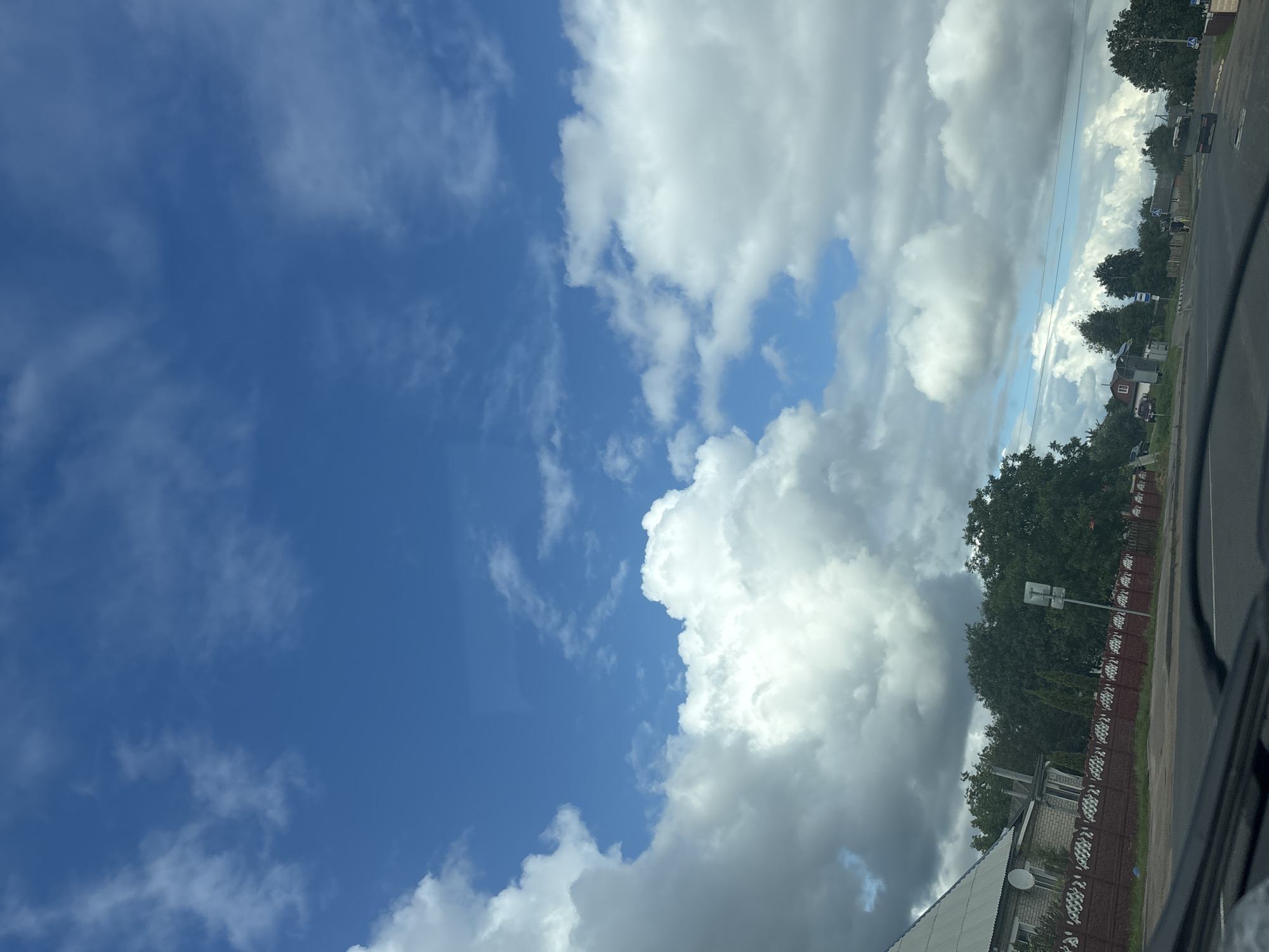 The image shows a rural landscape under a bright blue sky with scattered white clouds. In the foreground, the dashboard of a vehicle is visible, suggesting the photo was taken from inside a car. The middle ground features a road, a red fence, and several houses with trees behind them. The scene appears to be in a village setting. Based on the provided information, this scene is located in Kapyĺ, Belarus.