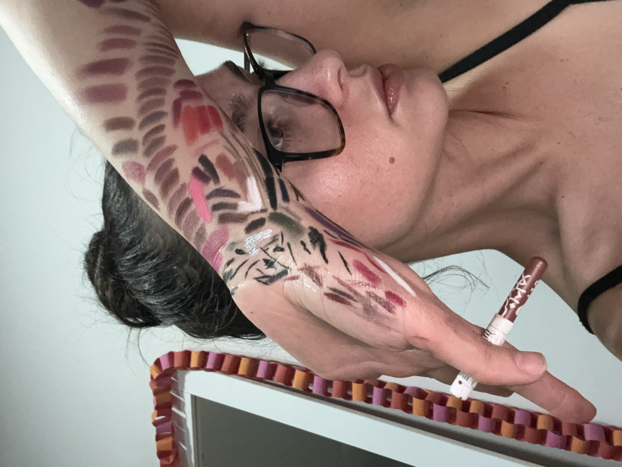 An individual, appearing to be an adult woman, is depicted in a close-up shot, angled so her head is tilted to the side, looking slightly upwards. She has dark hair, pulled back, and wears dark-framed eyeglasses. Her lips are slightly parted and have a glossy finish. A thin black strap, possibly from clothing, is visible on her shoulder.
Her left arm, from the upper arm down to her hand, is extensively covered with numerous makeup swatches and artistic applications. The upper arm features orderly rows of various shades, predominantly browns, mauves, and reds. On her forearm and hand, the makeup forms more abstract and expressive patterns, including strokes of pink, red, purple, black, and metallic silver/white, some resembling abstract art or scribbles. She holds a brown makeup pencil or crayon between the fingers of her left hand, which appears to be the tool used for the makeup application. Visible text on the pencil includes "XAI" or "YAI" and partially "BEAUTY".
In the foreground, a decorative paper chain, featuring segments in red, orange, and pink hues, is resting on a white, flat surface, possibly a table or shelf. The background is a plain white or light-colored wall. The overall scene suggests an activity of testing various makeup products or engaging in artistic makeup application on the skin. No specific time of day or emotional state is discernible. The location context provided is Krasnoyarsk, Russia, though no specific visual cues for this are present in the image.