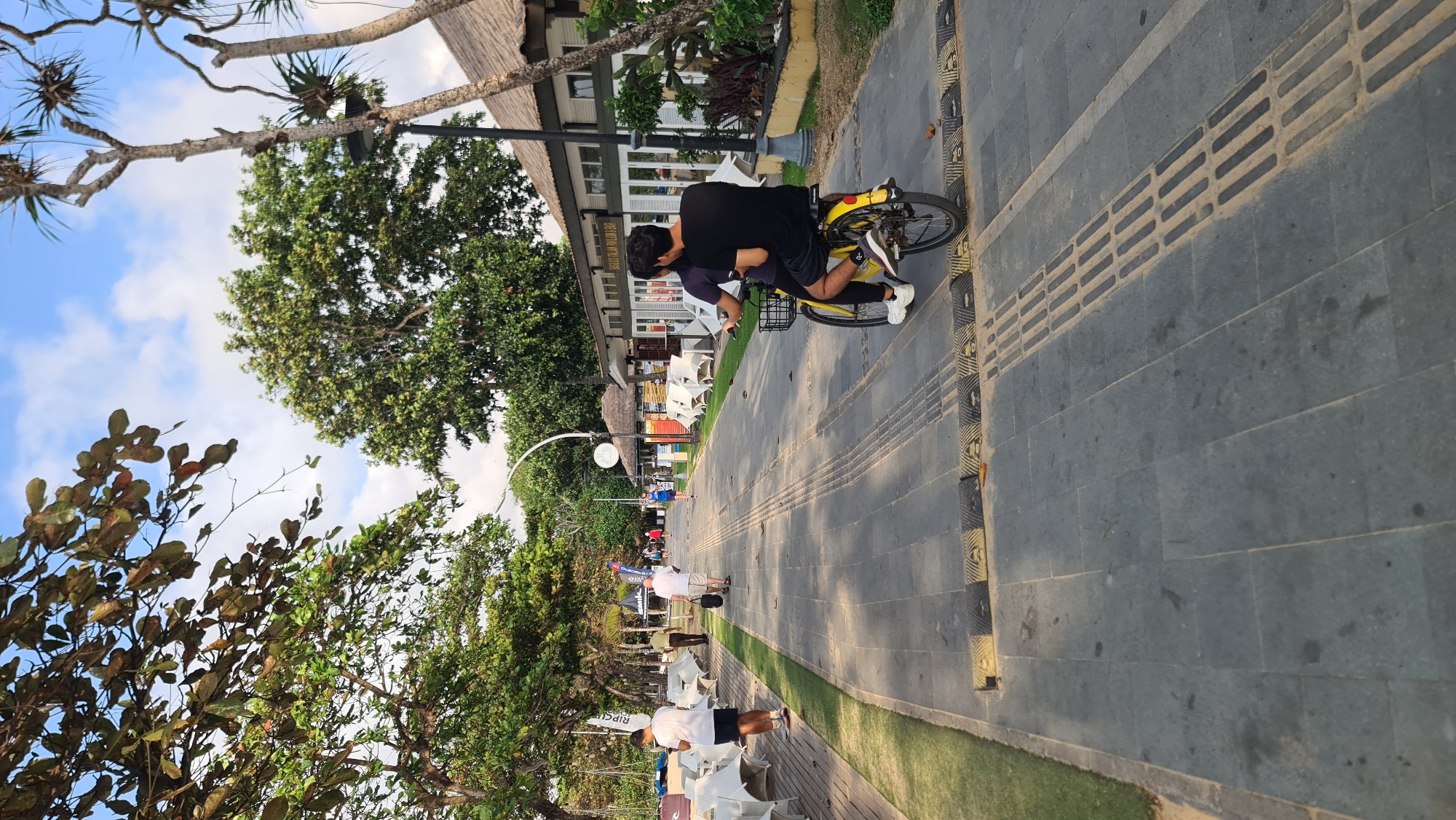 The image depicts a paved walkway alongside a beach in Denpasar, Indonesia, with a backdrop of lush trees and a few buildings that appear to be restaurants or cafes. The walkway is made of dark grey stones and features tactile paving for the visually impaired. In the foreground, two individuals are riding a yellow bicycle, with one person pedaling and the other seated behind. The person in front wears a dark t-shirt and shorts, while the person in the back wears a black t-shirt and shorts. Both are wearing sneakers. The bicycle has a basket on the front. The walkway is lined with speed bumps. To the left of the walkway, there is a grassy area with white chairs and tables, suggesting an outdoor seating area for a nearby establishment. Further down the walkway, several other people are walking, and some are visible in the background near the buildings. The sky is blue with scattered white clouds. The overall scene suggests a leisurely day in a coastal urban area.