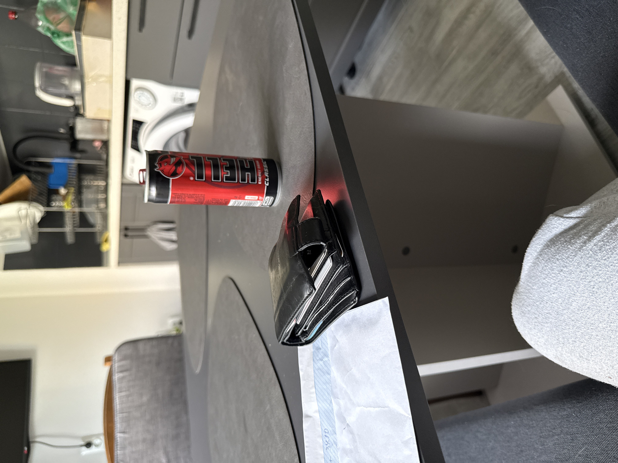 The image shows a can of Hell energy drink and a black wallet on a gray surface, likely a table. Part of a white washing machine is visible in the background, along with some kitchen equipment and cabinets. A gray chair and a leg covered in gray clothing are also in the frame. The location seems to be indoors, possibly a kitchen or dining area, but there is no information about whether the scene is in Stara Zagora, Bulgaria.
