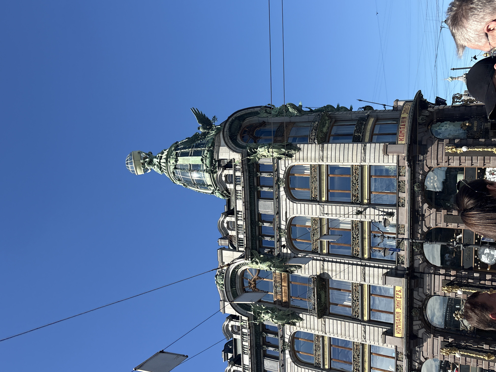 This image captures the iconic Singer House, also known as the Dom Knigi, located on Nevsky Prospekt in Saint Petersburg, Russia. The ornate Art Nouveau building, constructed between 1902 and 1904, is prominently featured against a clear blue sky. Its distinctive features include a glass dome topped with a globe and an eagle sculpture, as well as numerous statues and decorative elements adorning its facade. The building's signage, displaying "КОМПАНИЯ ЗИНГЕРЪ," is visible. Several people are present in the foreground, partially obscuring the lower portion of the building, suggesting it is a public area. Electrical wires crisscross the sky above the building, indicating an urban setting. The overall scene conveys a bright, sunny day in a historic city.