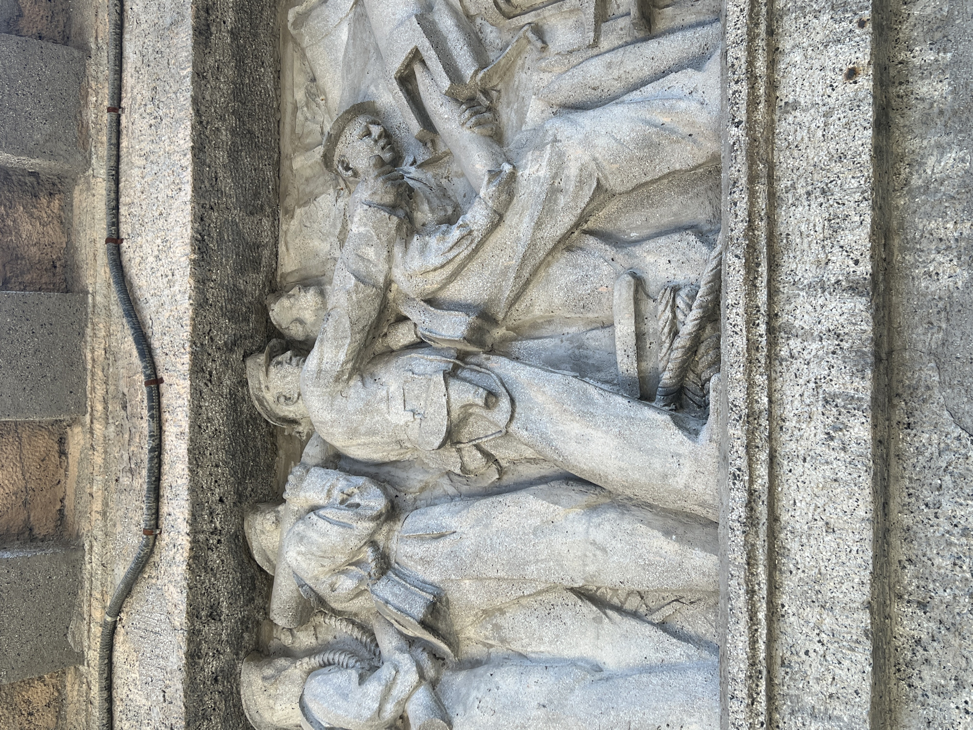 This image shows a grey, carved stone bas-relief sculpture set within a stone architectural element. The sculpture depicts several figures lying atop each other, possibly representing a group of fallen soldiers or laborers. One figure near the top appears to be holding a rectangular object overhead. The carving is highly detailed, showcasing clothing and facial features. The surrounding stone structure is textured and worn. The location is unknown.