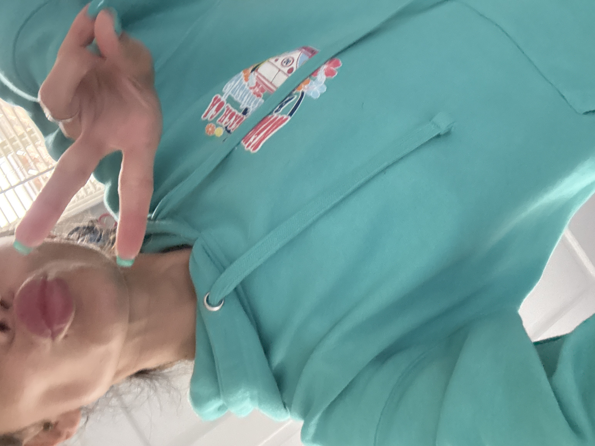 A casual, close-up image features a person wearing a vibrant teal or aqua-colored hooded sweatshirt. The person's head is visible in the lower left portion of the frame, with their tongue playfully extended from their mouth. Their left hand, positioned prominently in the upper left, displays a peace sign (V-sign) towards the viewer. A simple ring adorns their ring finger, and their fingernails are painted with a light blue-green polish.
The hooded sweatshirt features a colorful graphic design on its chest. The design appears to be a stylized, whimsical depiction of beach-related elements, possibly including a surf van or bus, and another abstract shape resembling a surfboard or flag. The graphic utilizes bright colors such as reds, whites, blues, yellows, and oranges. The hoodie also shows its matching teal drawstrings with shiny silver eyelets.
The background is largely out of focus and appears to be a light, indistinct color, suggesting an indoor setting. There are faint hints of a grid-like structure, possibly a window or vent, in the blurred background behind the hand. The overall impression is one of playfulness and casualness, captured in a candid moment. The unique angle of the shot, which is rotated 90 degrees clockwise, contributes to its informal and personal feel. No discernible text is clearly readable within the graphic or elsewhere in the image.
