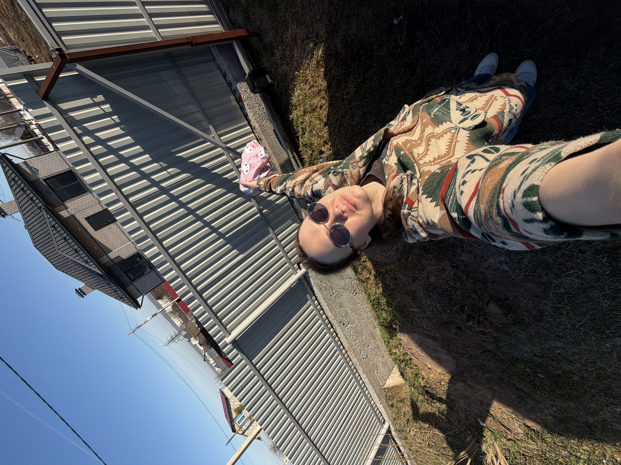 A person lies on the ground near a metal fence and metal-roofed structure, seemingly taking a selfie. The ground is a mix of dry grass and dirt. The person is wearing sunglasses and a patterned shirt. In the background is a house and a clear blue sky. It is daytime and sunny. The location is likely Chaikovsky, Russia.