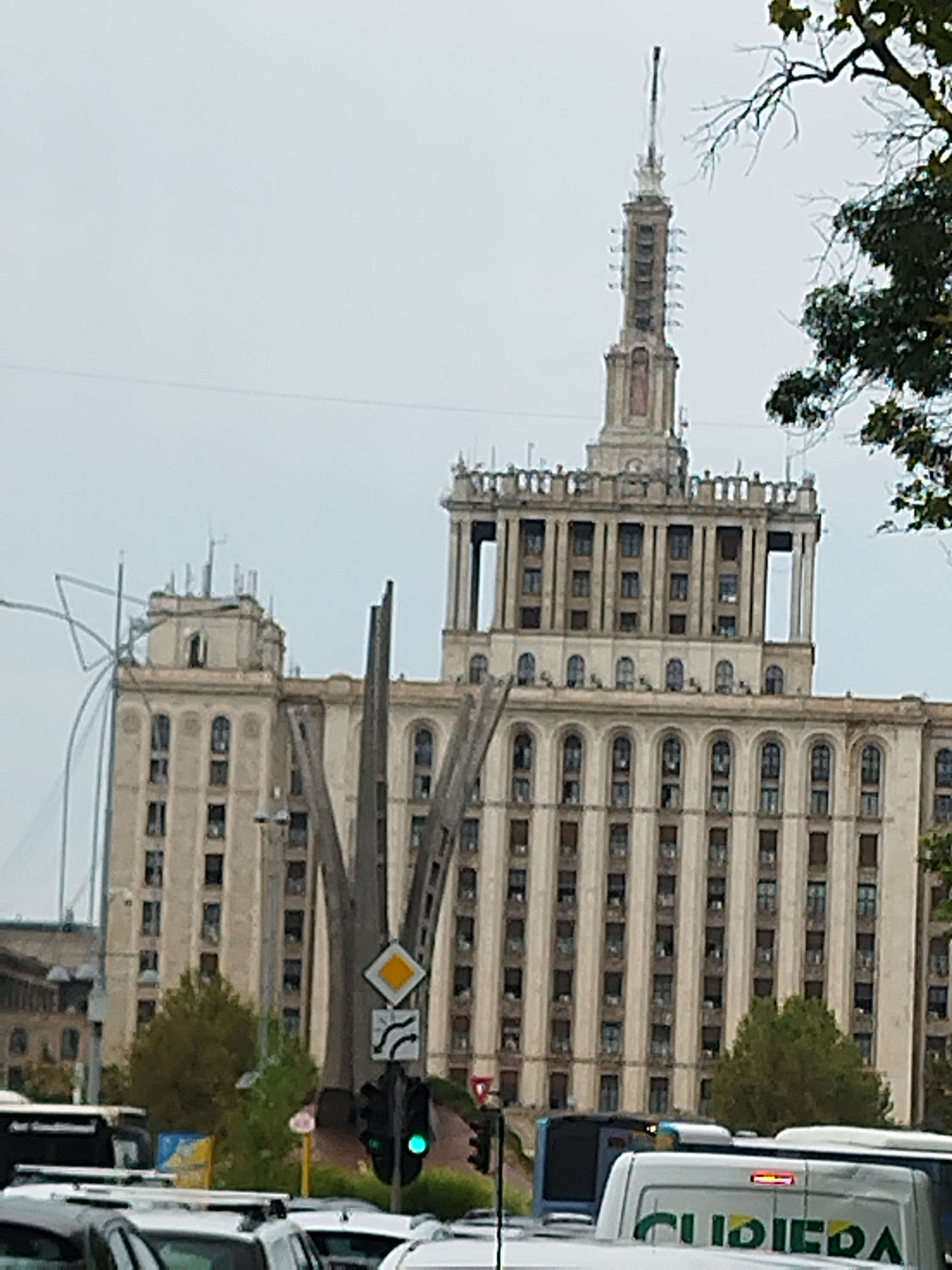 The image shows the House of the Free Press, a landmark building in Bucharest, Romania, on an overcast day. The Brutalist skyscraper, characterized by its imposing facade with numerous repetitive windows and ornate architectural elements, dominates the scene. In the foreground, traffic lights show a green light, indicating movement. Several vehicles, including cars and a bus with the logo "CURIEPA" visible, are present on the street. Trees with autumnal foliage are scattered around the base of the building and in the mid-ground. A tall, abstract sculpture made of metal is positioned in front of the building. The sky is a uniform grey, suggesting a cloudy or possibly rainy day. The overall impression is an urban landscape with a prominent architectural monument and daily city life unfolding around it.