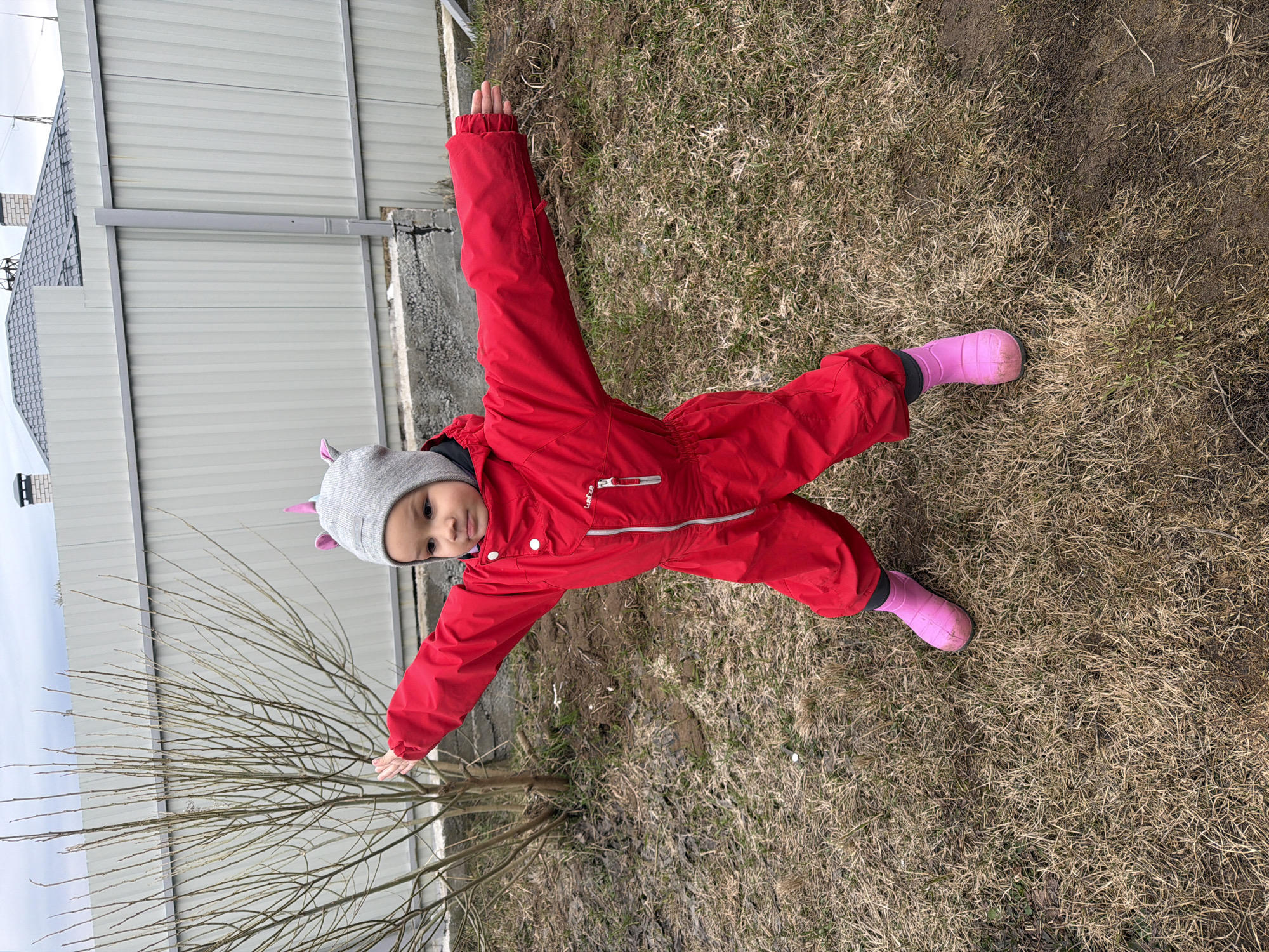 A child in a red snowsuit and pink boots lies on their back on a patch of dry, brown grass. Their arms are outstretched to their sides, forming a cross shape with their body. They are wearing a grey hat with small pink protrusions, likely meant to resemble animal ears. A bare-branched tree is visible to the left of the child. In the background, there is a corrugated metal fence, and a house with a grey roof. The picture was taken outside, likely in the city of Chaikovsky, Russia.