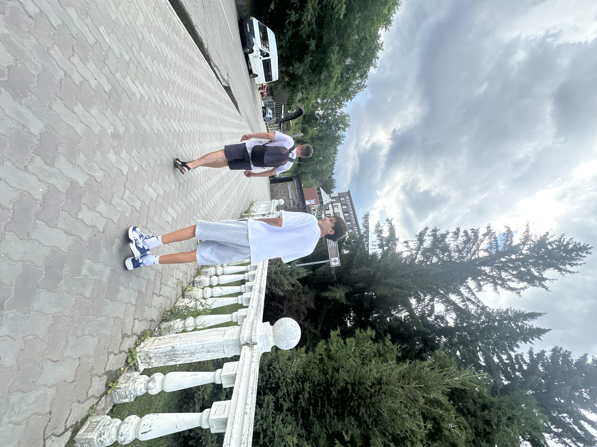 In Belokurikha, Russia, two individuals are walking away from the camera on a paved walkway lined with a white ornate railing. The person in the foreground is a young male wearing a white t-shirt, grey shorts, white socks, and blue and white sneakers. The person behind him is an adult male wearing a grey t-shirt, dark shorts, and sandals, carrying a grey and red backpack. A white van and other vehicles are visible on a road to the left of the walkway. Tall evergreen trees and a multi-story building with signage are situated behind the walkway and railing. The sky is overcast with dramatic clouds. The scene appears to be a public area, possibly a resort or a scenic route within the city.