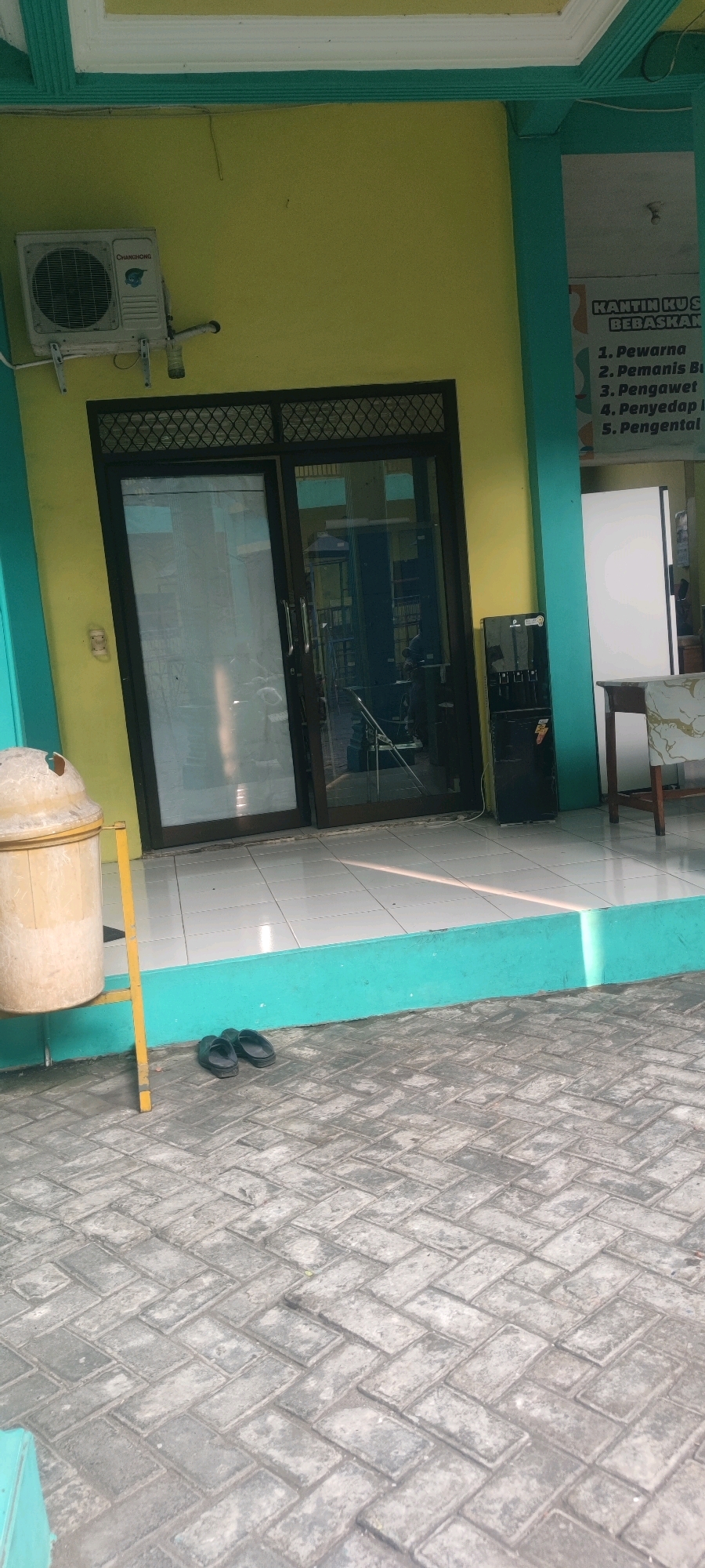 The image shows the exterior of a building in Banjarsari, Indonesia. The building has a bright yellow facade with teal trim and a white overhang. There is a glass door with dark frames in the center of the building. To the left of the door, an air conditioning unit is mounted on the wall. To the right of the door, a black water dispenser is visible. A portion of a sign with Indonesian text is visible on the right side of the building. In front of the building, the ground is paved with grey interlocking bricks. To the left of the door and near the edge of the image, a light-colored trash can on a yellow stand is present. A pair of black flip-flops are on the pavement in front of the teal trim at the base of the building. The scene appears to be outdoors in daylight, with shadows cast on the ground.