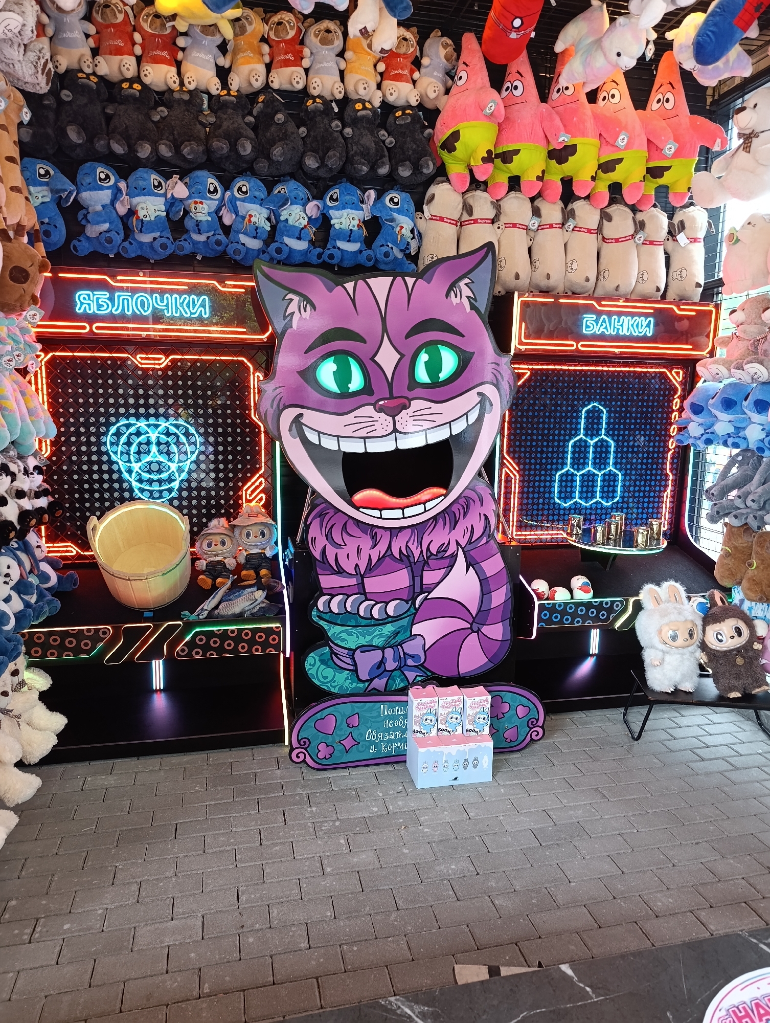 A large, cut-out cardboard figure of the Cheshire Cat from Alice in Wonderland is the central focus of the image. The cat is depicted with a wide, grinning mouth, green eyes, and purple and pink striped fur. It is standing in front of a game booth that features neon lights and plush toys. The game booth has two sections, one labeled "ЯБЛОЧКИ" (Apples) and the other "БАНКИ" (Jars). The "Apples" section has a large wicker basket and two small plush toys dressed as farmers. The "Jars" section has a rack of small cups and a honeycomb-like structure outlined in neon blue. Plush toys of various characters, including Stitch, Patrick Star from SpongeBob SquarePants, and teddy bears, are displayed on shelves above and around the game booth. In the foreground, there are a few more plush toys and some boxes. The ground is paved with gray bricks. The scene appears to be an outdoor amusement or carnival setting. The image was taken in Kaliningrad, Russia.