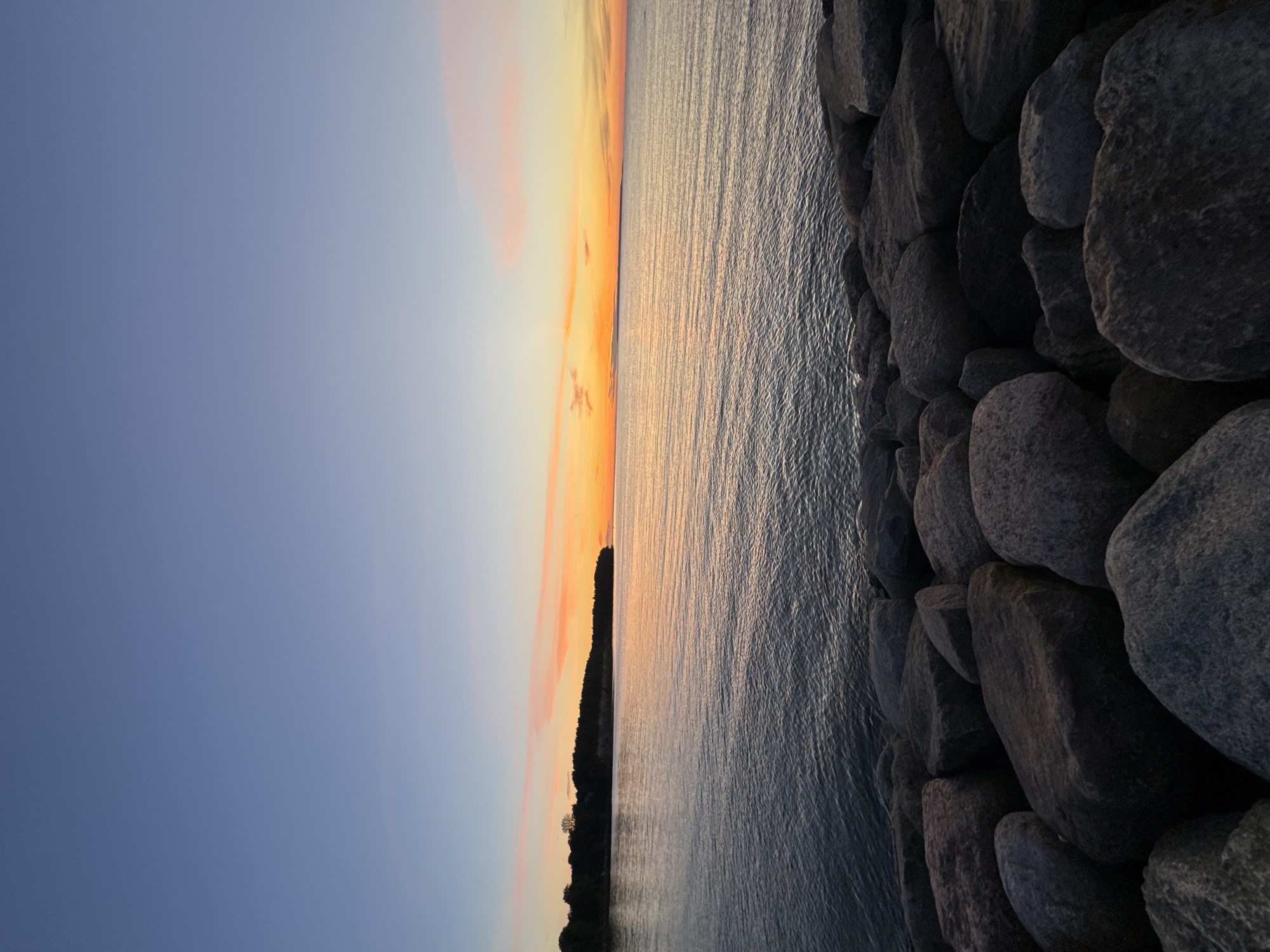 The image captures a serene coastal scene at dusk, likely along the shores of Tallinn, Estonia. In the foreground, large, dark, rounded rocks are piled along the edge of the water, creating a natural barrier. The water itself is calm, with gentle ripples reflecting the soft, warm colors of the sunset. Beyond the water, a densely wooded shoreline forms a dark silhouette against the horizon. The sky above transitions from a deep blue at the top to shades of orange, pink, and yellow near the horizon, indicating the sun has recently set or is about to set. There are no people or animals visible in the frame. The overall impression is one of natural beauty and tranquility.
