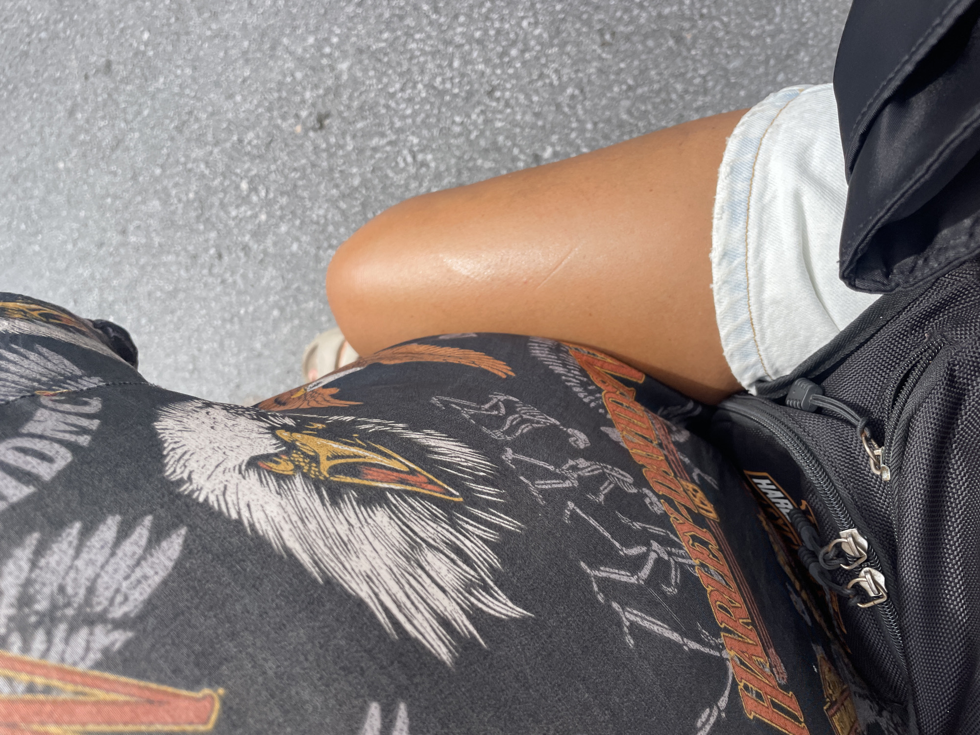 The image shows a close-up of a person's lap. The person is wearing light-wash denim shorts and a dark-colored shirt with a Harley Davidson eagle graphic and skeletons. A black backpack rests on the person's lap. The background appears to be asphalt. The image was likely taken in Choeng Thale, Thailand.