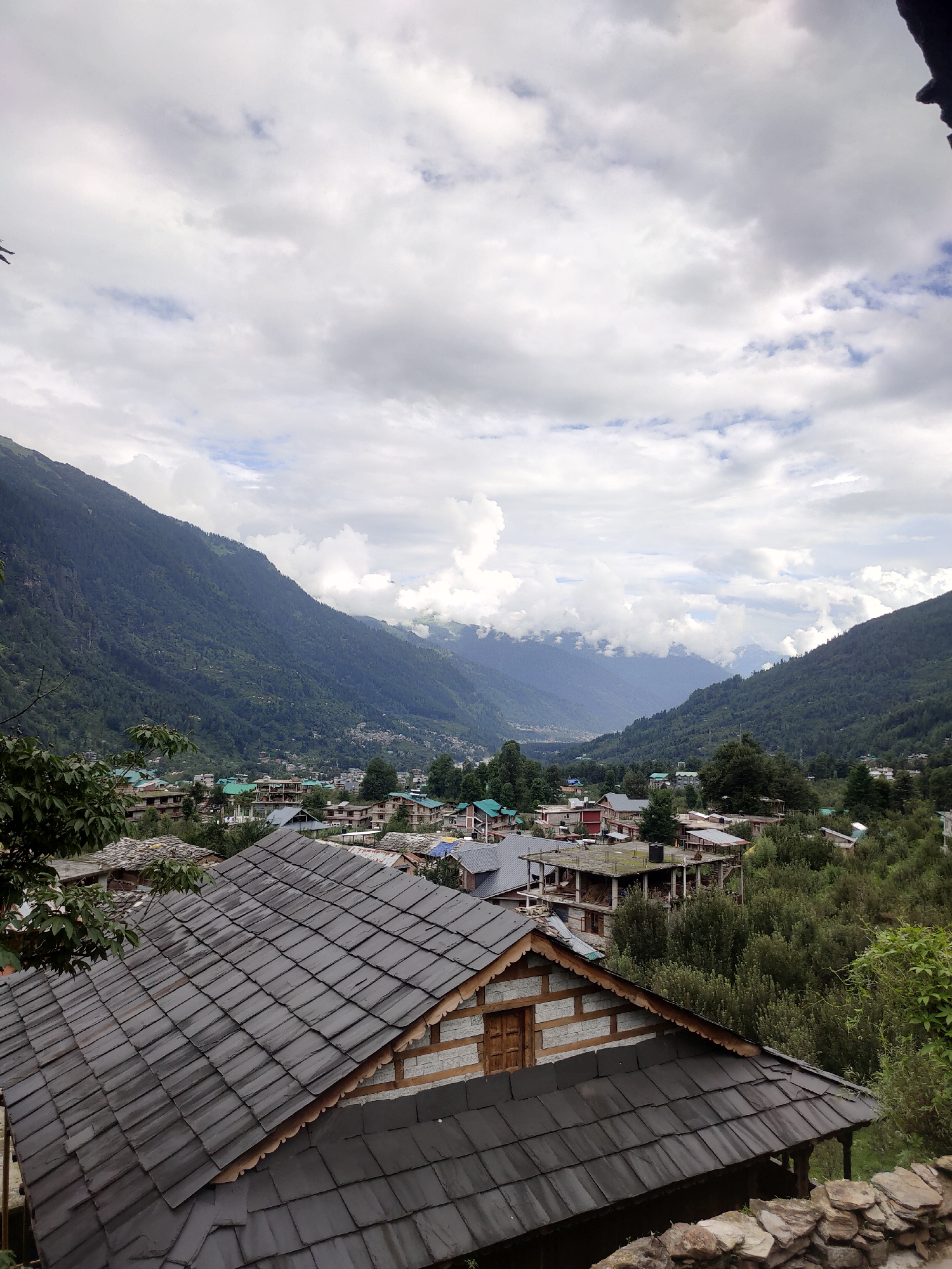 The image depicts a panoramic view of the village of Burwa, India, nestled in a valley surrounded by lush green mountains. The foreground is dominated by the dark, tiled roof of a traditional wooden and stone house. Beyond this, the village sprawls with more houses, many featuring similar tiled roofs and concrete structures. Buildings are interspersed with dense green trees and foliage. In the background, majestic mountains rise, their slopes covered in dark green vegetation. The sky is overcast with dramatic, billowy clouds, with patches of blue sky visible. The scene conveys a sense of rural tranquility and the raw beauty of a mountainous landscape.
