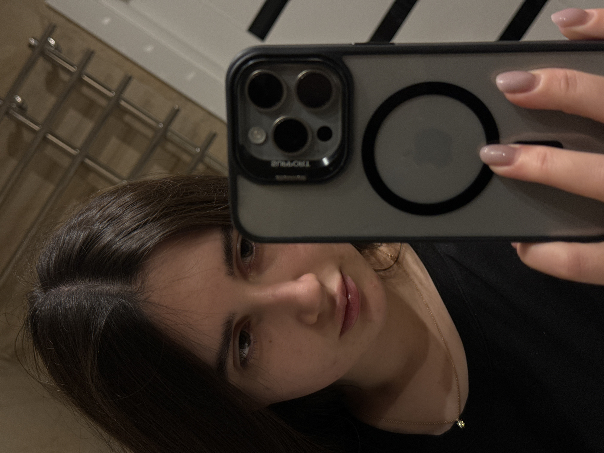 This media file captures a selfie of a young woman with dark hair and light skin, taken in what appears to be an indoor setting, possibly a bathroom given the presence of a towel rack in the background. The woman is looking at the camera, with a slight smile. She is wearing a black top and a delicate gold necklace with a small pendant.
The photograph is taken from a slightly angled perspective, with the woman holding a smartphone in her hand to take the picture. The phone case is visible, showing a triple camera system and the Apple logo. The woman's hands, with neatly manicured nails painted in a muted pinkish-beige color, are also visible, resting on the phone.
The lighting suggests it is daytime, but no specific weather conditions are discernible. The background is somewhat blurred, but the distinct structure of a chrome towel rack indicates a domestic environment. There is some faint text visible on the phone case, appearing to be "TROCHIL" or similar, though it is partially obscured. The overall mood of the image is casual and relaxed. The location context provided is Vinnytsia, Ukraine.
