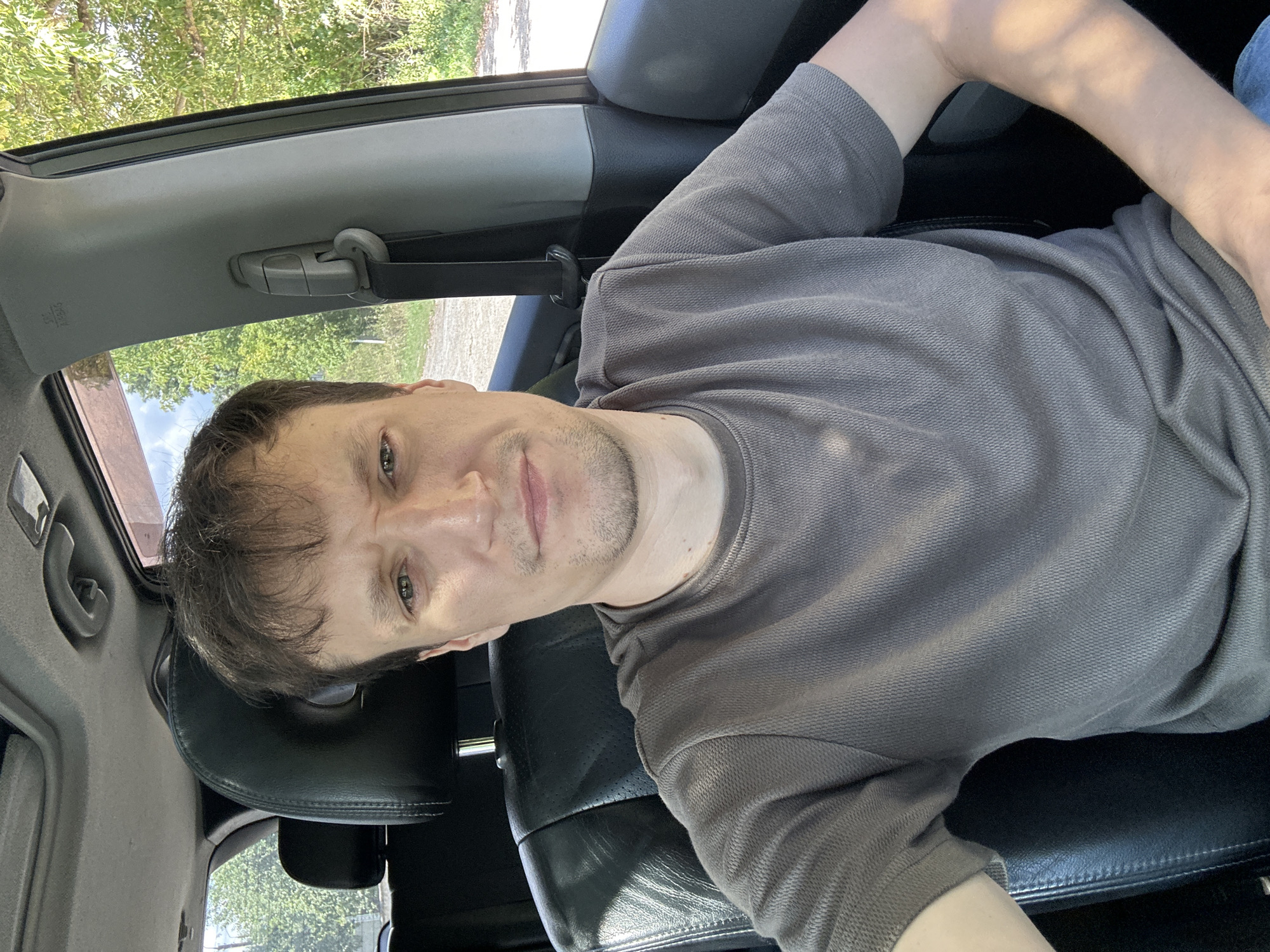 The image is a selfie of a man in a car, taken from the chest up. He is sitting in the driver's seat, wearing a grey short-sleeved shirt. The car's interior is visible, including a black leather seat and part of the dashboard. Through the car window, trees and a dirt road can be seen, suggesting an outdoor, possibly rural, location. The lighting indicates it is daytime. The man's expression is neutral. There are no specific landmarks visible that would pinpoint the location to Makiv, Ukraine.