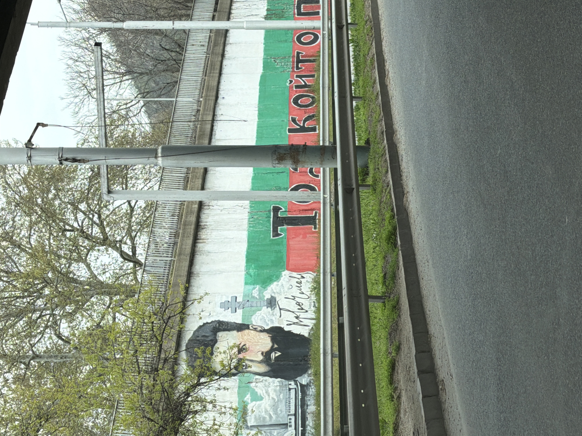 The image captures a mural painted on a wall alongside a road in Plovdiv, Bulgaria. The mural depicts Vasil Levski, a Bulgarian revolutionary and national hero, against the backdrop of the Bulgarian flag. Alongside the portrait and flag is the phrase "Това Който". Trees with budding leaves are present in the foreground, partially obscuring the mural. A metal guardrail separates the mural from the road. The road surface is visible on the right side of the image. A metal post and railing appear to be partially obscuring the mural.