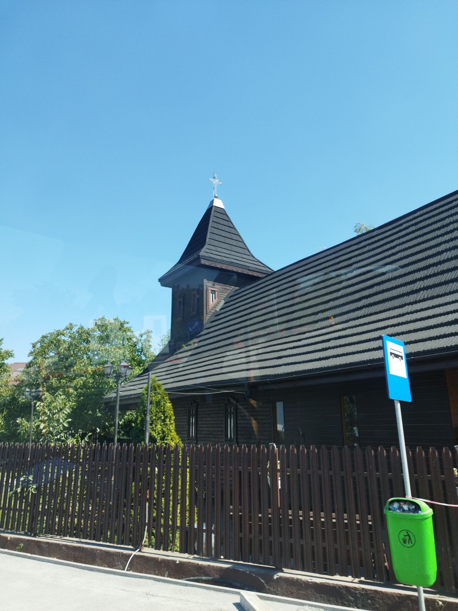The image depicts a wooden church with a prominent spire topped with a cross, located in Buftea, Romania. The church has a dark-colored, shingled roof and dark wooden walls with visible window frames. In front of the church, there is a brown wooden fence. To the right of the church, a blue bus stop sign is visible, with a green trash bin situated next to its pole. The sky is clear and blue. The scene appears to be captured from a moving vehicle, as suggested by reflections on the church's windows.