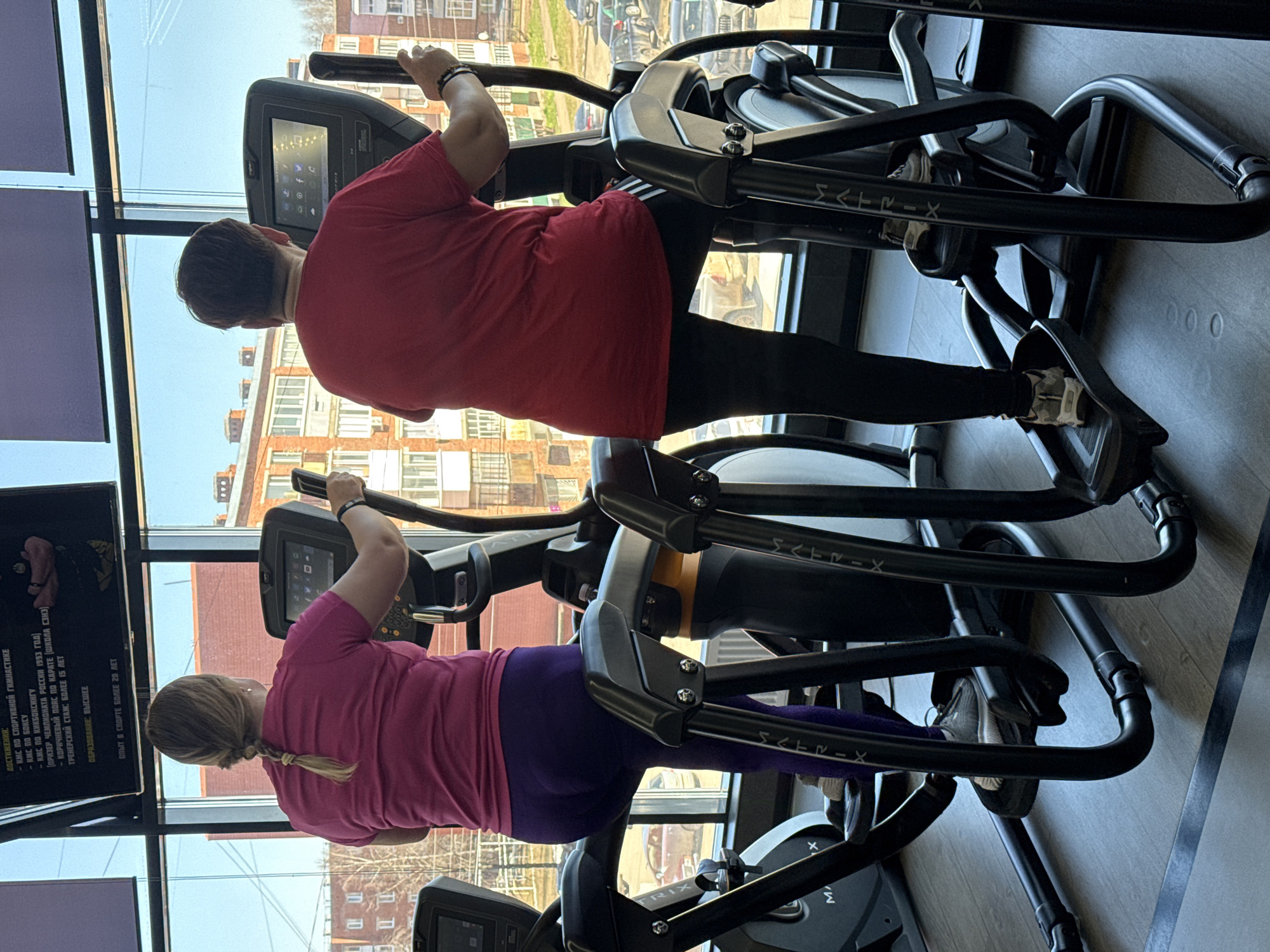 Two individuals are using elliptical machines in a fitness center. The person in the foreground wears a pink shirt and purple leggings, while the other person is clad in a red shirt and black pants. Large windows behind the exercisers reveal buildings, possibly in Votkinsk, Russia. In the bottom right of the frame, a grey colored wooden floor can be seen. The ellipticals are black and branded with "Matrix". A television screen with text in Russian is visible on the left. The machines have control panels.