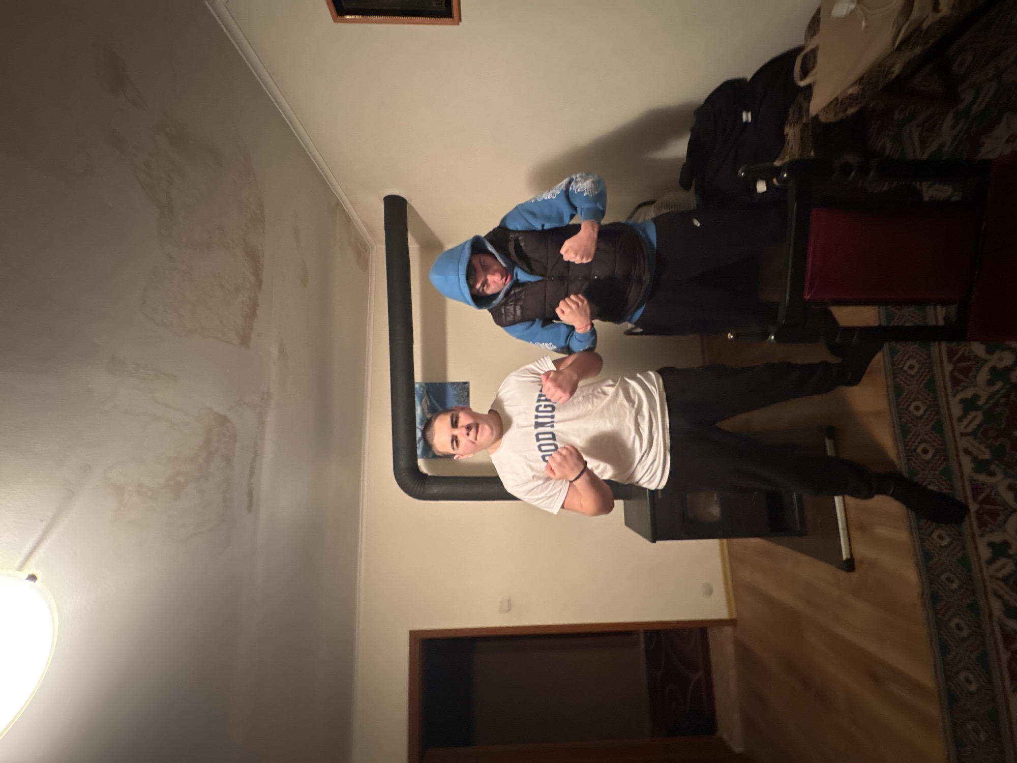 Two men are posing indoors. The man in the foreground is wearing a white t-shirt and black pants, while the man in the background is wearing a blue hoodie and a black vest. Both are clenching their fists. Behind them is a black stovepipe. The room has a beige wall with what appears to be water damage on the ceiling. There is a dark wood cabinet in the background. The location is Unknown, Unknown.