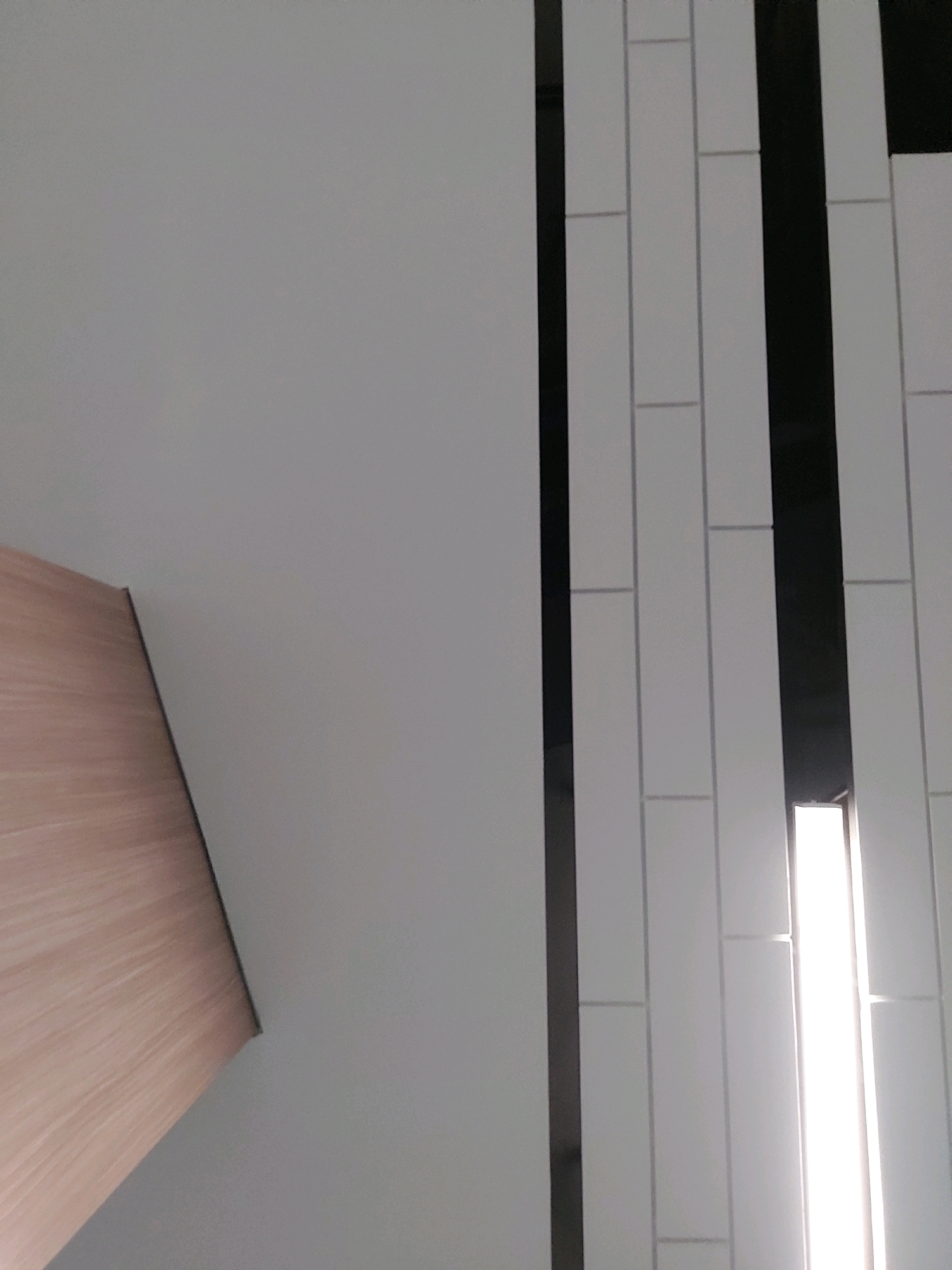 The image shows a close-up view of a wall with a wood-paneled section on the left and a tiled section on the right. The wood paneling is light brown with a visible grain pattern. The tiled section consists of vertical white tiles with thin black grout lines. There is a vertical black stripe separating the wood paneling from the tiled section. A bright light source is visible within the tiled section on the far right, suggesting a light fixture. The image appears to be taken indoors, possibly in a modern interior space. The location in Bekasi, Indonesia is not identifiable from the visual information provided.