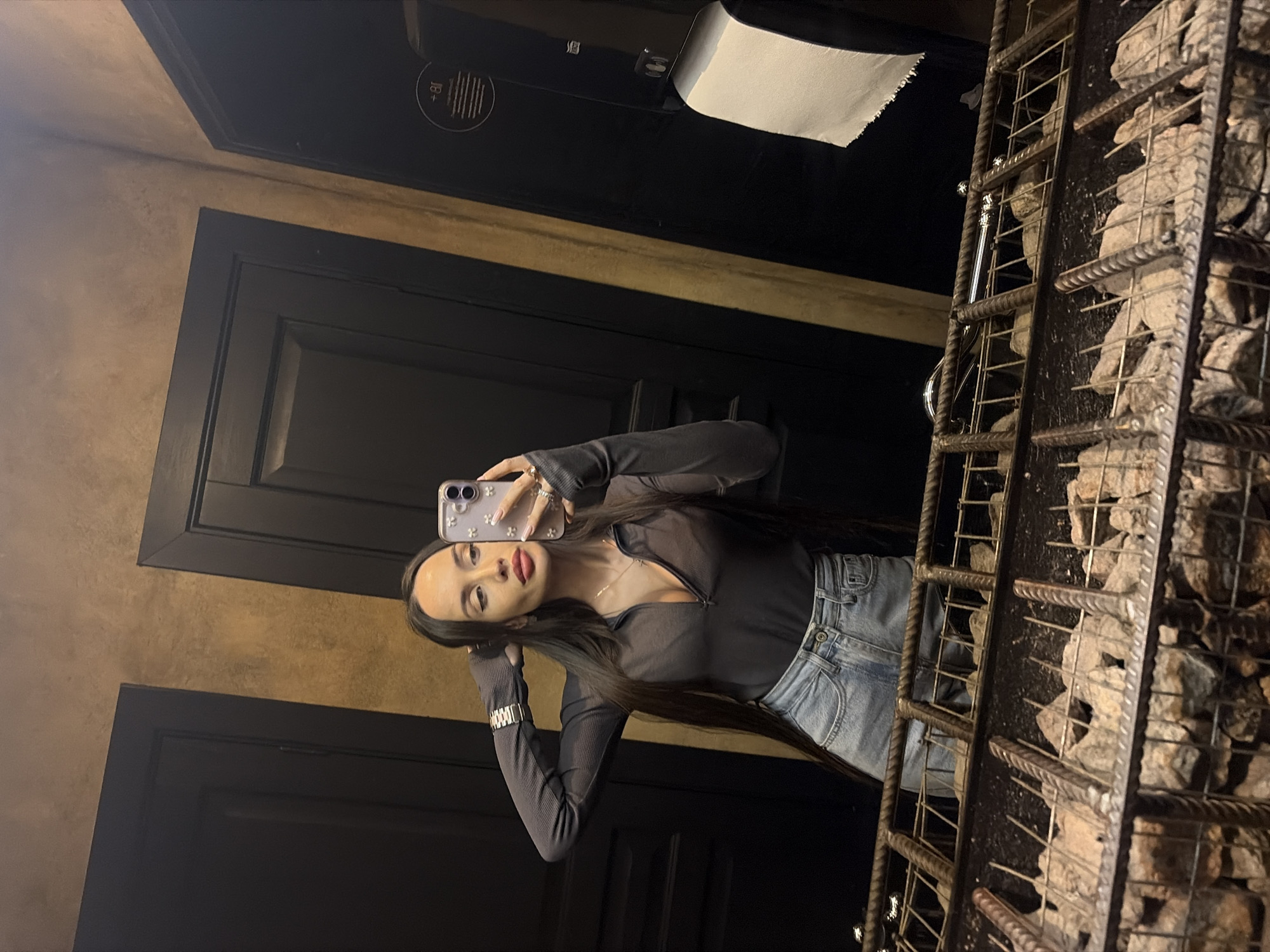 A young woman is taking a selfie in what appears to be a bathroom or changing room. She is holding her phone in her left hand, the screen facing the viewer. The woman is wearing a dark long-sleeved top with a zippered neckline and light-wash jeans. She has long, dark hair and is wearing jewelry, including a watch and a necklace.
The background consists of a dark-colored door and a wall with a textured, potentially painted surface. To the right of the woman, there is a structure constructed from metal bars filled with rocks, possibly a decorative element. Above the door, a toilet paper dispenser is visible.
The lighting in the room is dim, creating shadows and adding contrast to the image.
Based on the details visible, it is impossible to determine the location of the bathroom with certainty. Therefore, I can't provide confirmation if the bathroom is in Saint Petersburg, Russia.
