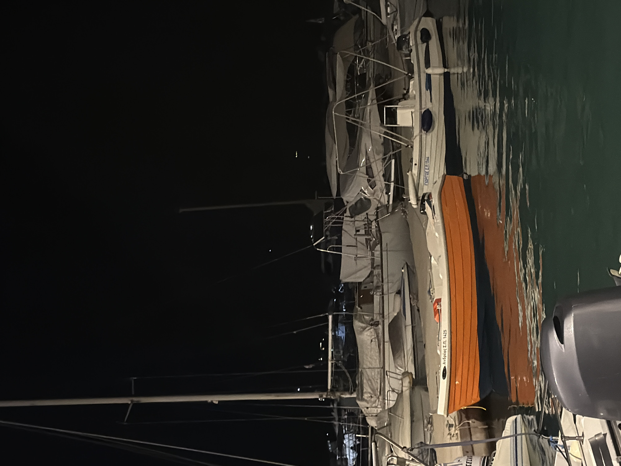 The image depicts a nighttime scene at a marina in the Municipal Unit of Alyzia, Greece. The foreground is dominated by the dark, calm water of the harbor. Several boats are moored, their outlines visible against the blackness.
In the mid-ground, a vibrant orange and blue rowboat is the most prominent subject, floating on the water. It has some white text on its side. Adjacent to it, a white motorboat with blue trim is also docked. Behind these, a larger sailboat with a white hull and mast is partially visible, covered with a gray tarp. Further in the background, more boats, some covered and some with their structures visible, are lined up. Distant lights from what appears to be the shore or other structures dot the dark horizon. The overall atmosphere is still and quiet, typical of a marina at night.
