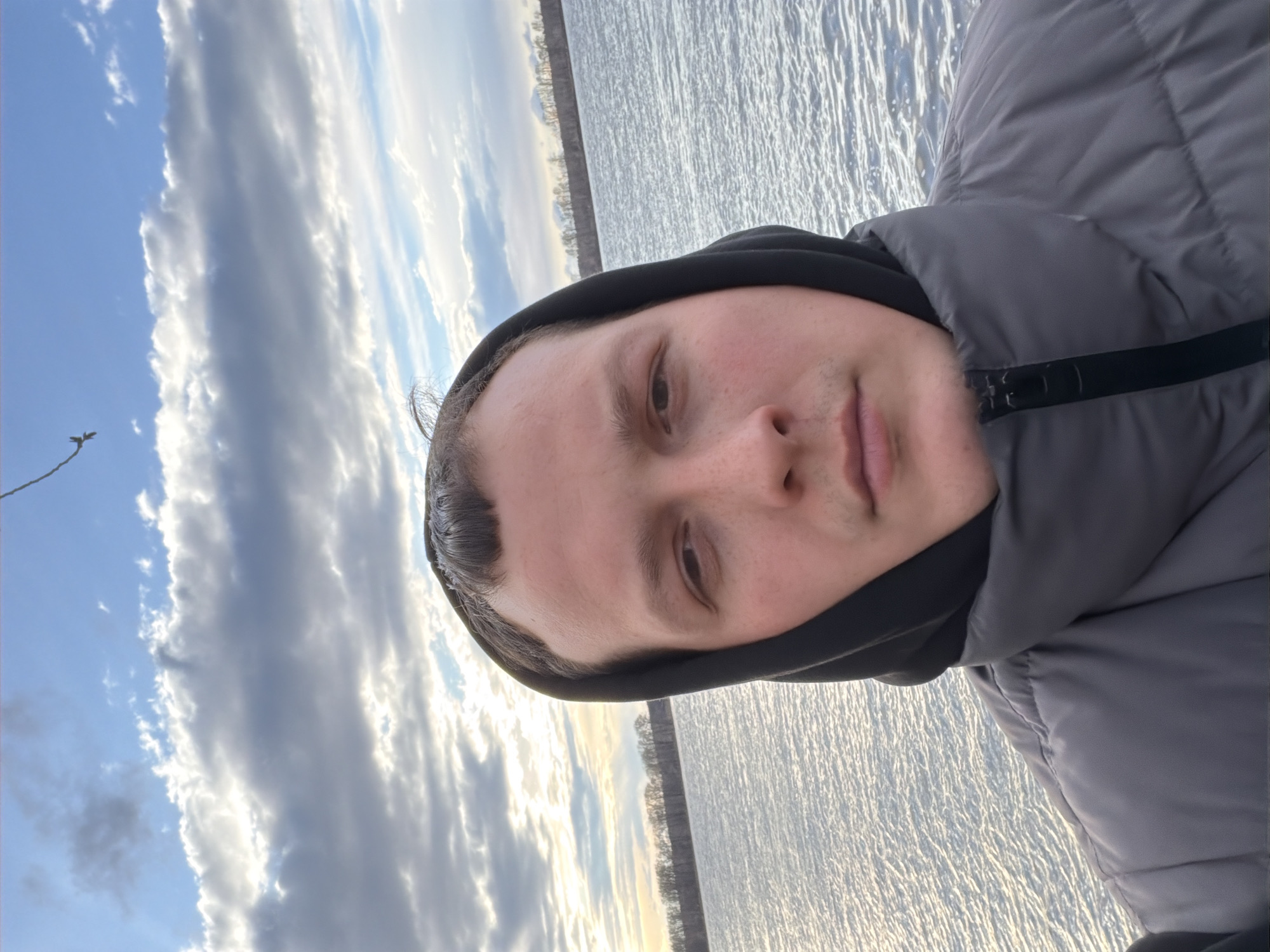 A young adult male is featured in a close-up selfie, taken outdoors in Chaikovsky, Russia. He looks directly forward with a neutral to slightly pensive expression. He is wearing a black hooded sweatshirt under a grey puffer jacket, suggesting cool weather.
The background prominently displays a wide expanse of water, likely the Kama River given the location context. The water's surface is heavily textured with ripples or small waves, brightly reflecting the sky's light, almost appearing silvery. Beyond the water, a distant treeline, appearing bare, marks the horizon under a partly cloudy sky. The sky features patches of blue alongside long, streaky white and grey clouds, possibly stratocumulus or altocumulus.
The soft, diffused lighting suggests the photo was taken during either early morning or late afternoon. The overall scene depicts a calm, cool day by a large body of water. There is no visible text in the image.
