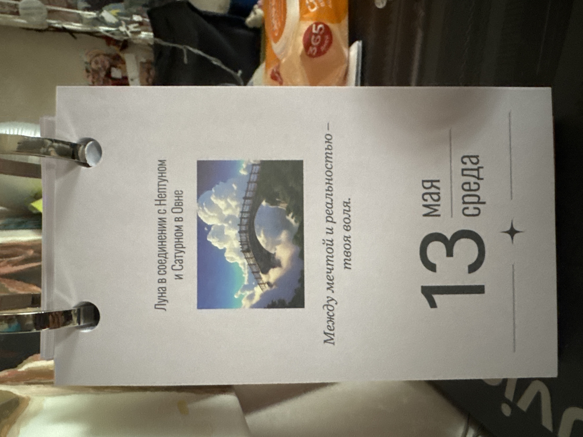 A photograph shows a flip calendar, oriented vertically in the frame. The page displayed is for May 13th, Wednesday ("среда" in Russian). The page includes a small image of a cloudy sky above a bridge. Text in Russian on the page reads "Луна в соединении с Нептуном и Сатурном в Овне," which translates to "Moon in conjunction with Neptune and Saturn in Aries." There is also another quote written in Russian: "Между мечтой и реальностью – твоя воля," meaning, "Between dream and reality is your will." The calendar is bound by metal rings. A product from the brand "365" is visible partially at the top of the frame. The calendar sits in a cluttered environment. There are no people visible in the photo. The location is unknown.