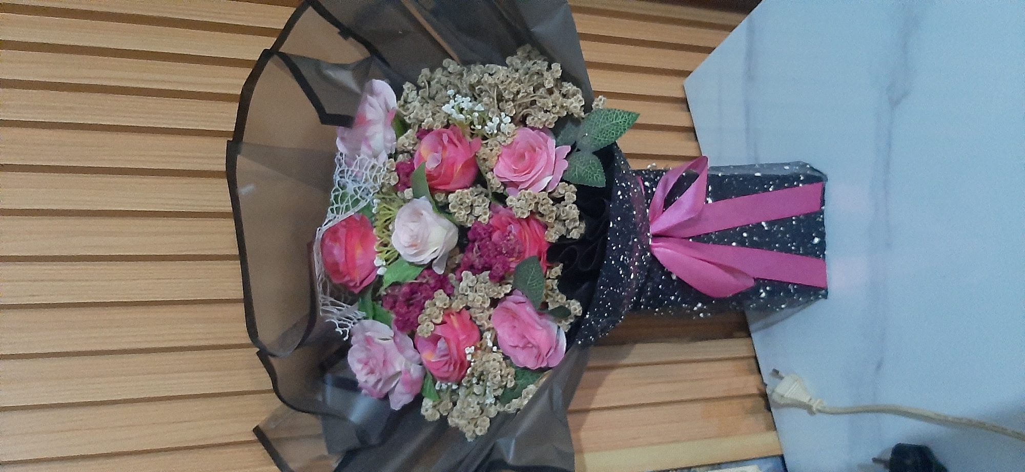 A bouquet of artificial flowers, predominantly pink and red roses, with small green leaves and brown filler flowers, is displayed in a black, glitter-covered vase. A bright pink satin ribbon is tied in a bow around the vase. The bouquet is wrapped in translucent black paper. The background consists of vertical wooden blinds. The vase sits on a white, marble-patterned surface. A light-colored electrical cord with a plug is visible on the left side of the image. The scene appears to be indoors, likely in a domestic setting. The overall impression is a decorative arrangement. The image does not provide specific details that would allow for pinpointing a location within Jambi, Indonesia.