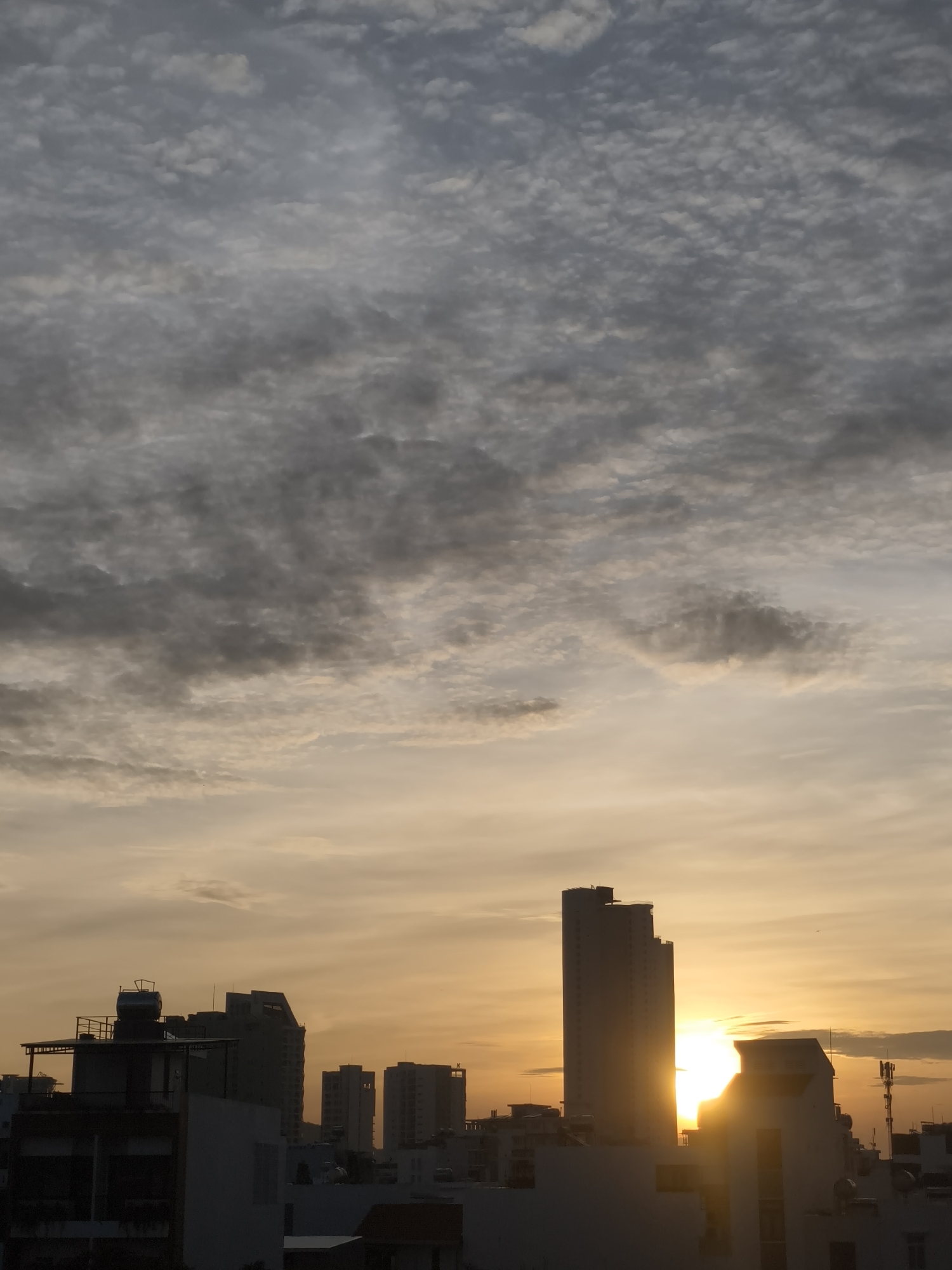 The image depicts a city skyline at sunset. The sky is filled with a mix of grey and white clouds, with a warm orange glow emanating from the setting sun on the right side of the frame. Silhouetted against the sky are several buildings of varying heights, forming a dense urban landscape. The buildings on the left are darker and appear to be closer, with visible rooftops and structural details. The sun is partially obscured by a tall, slender building in the center-right of the image, casting a bright glare. In the foreground, the tops of other buildings are visible, also silhouetted against the dramatic sky. The overall scene is one of a bustling city transitioning from day to night, with the warm hues of sunset creating a striking contrast with the dark silhouettes of the urban structures.