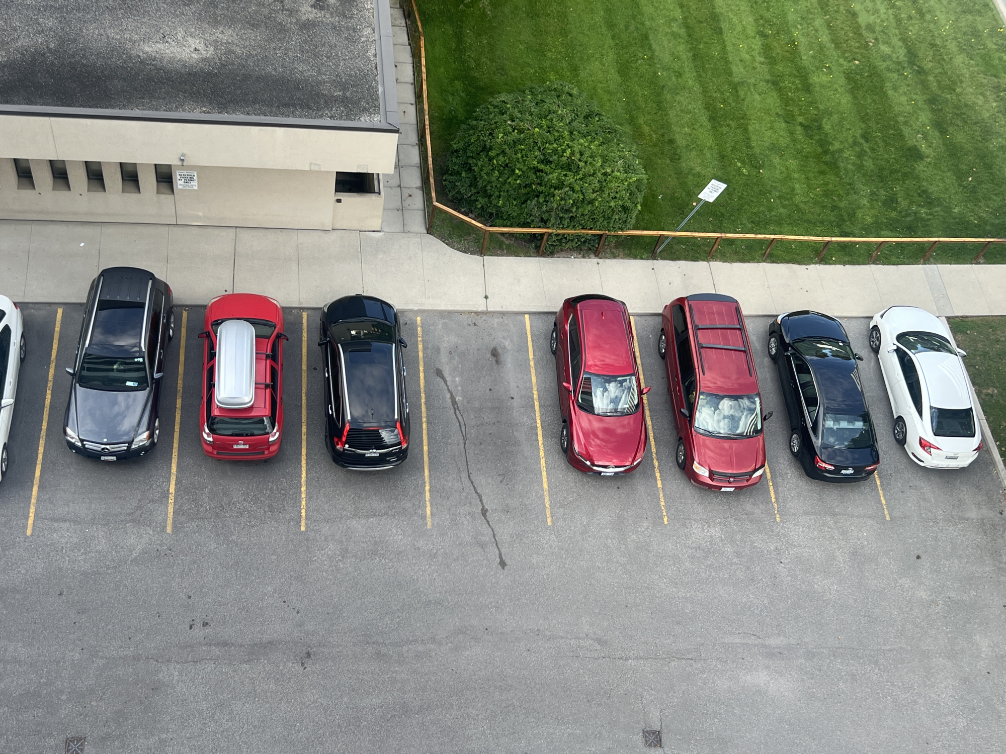 This is an aerial, full shot of a parking lot adjacent to a building and a grassy area. Nine cars are parked in marked spots. From left to right, the cars are a white sedan, a dark gray sedan, a red SUV, a black sedan, a red minivan, a black sedan, a black SUV, a red minivan, and a white sedan. A small white sign reading "RESERVED PARKING BY PERMIT" is visible attached to the building. The environment appears to be an outdoor urban setting, possibly a business park or residential complex. The city of Markham, Canada, is identified as the location.